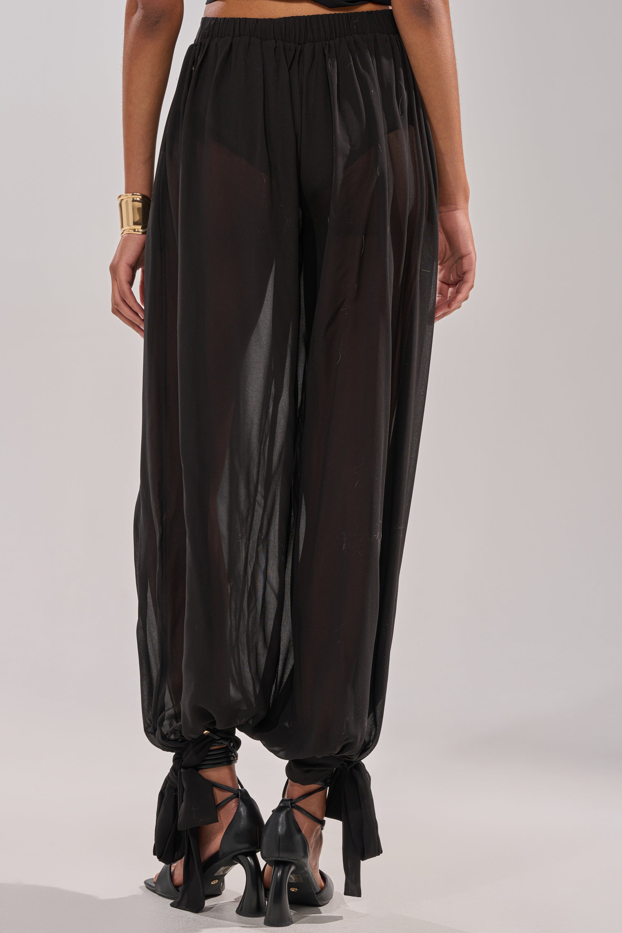 A person wears the TRENDY BALLON PANT: high-rise chiffon pants with an elastic waistband, wide legs gathered at the ankles and tied with bows. Sheer fabric reveals black underwear, styled with black heels and a gold bracelet for a chic look. 7