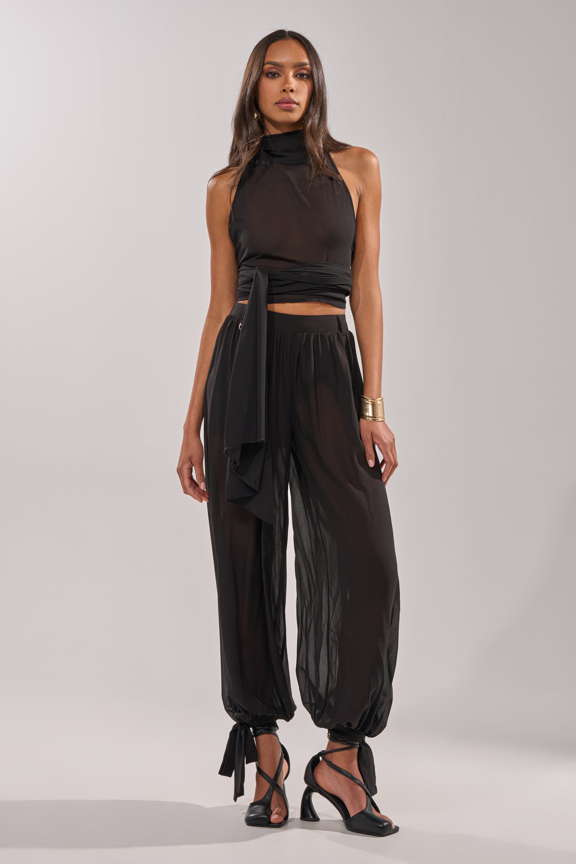 A woman models the TRENDY BALLON PANT, featuring ankle ties, paired with a sheer black halter top, black heels, and a gold cuff bracelet, set against a plain light gray background. 8