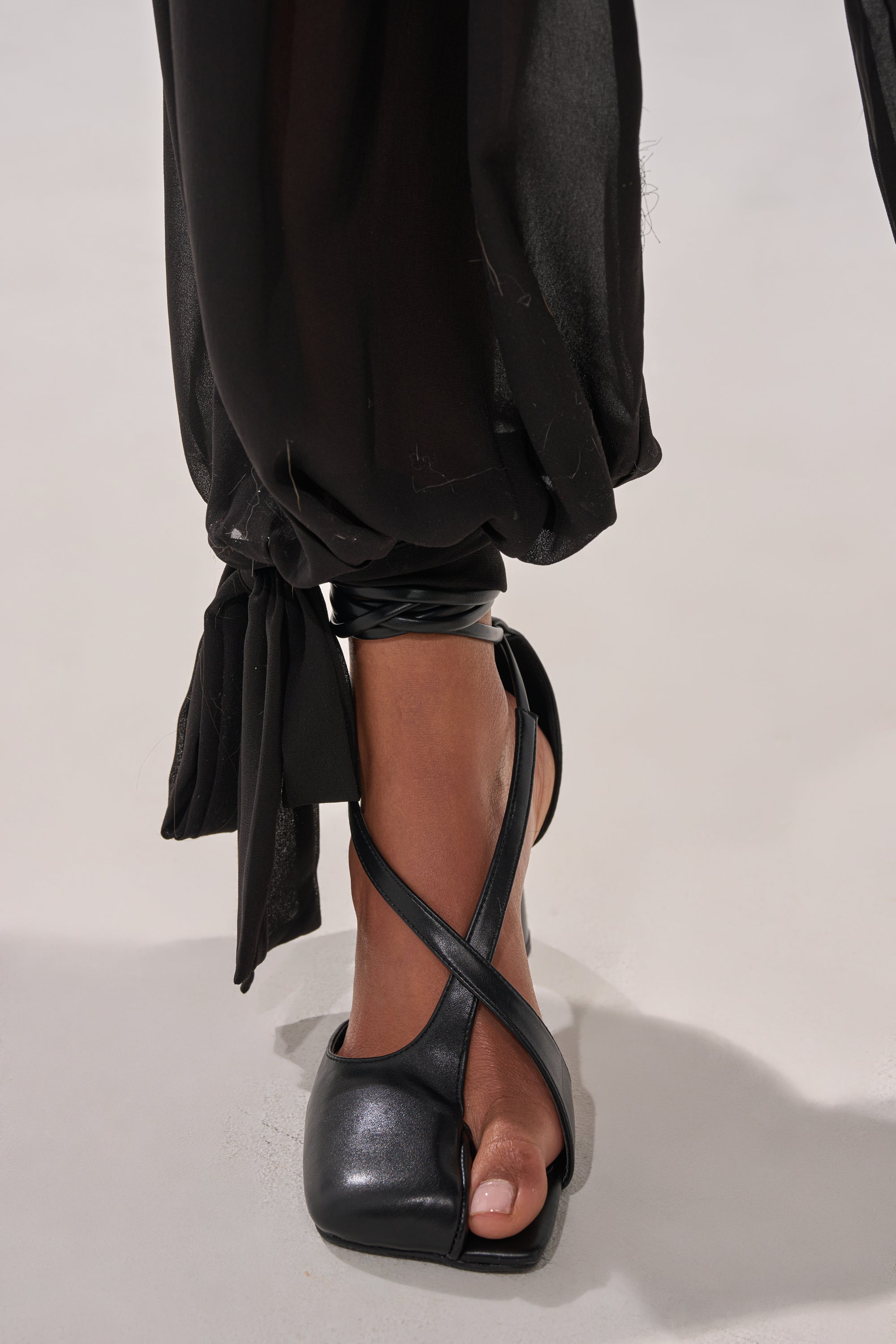 A close-up of a person wearing black crisscross open-toe heels and TRENDY BALLON PANT with sheer black chiffon fabric and tied ankles, walking on a light-colored surface. 9