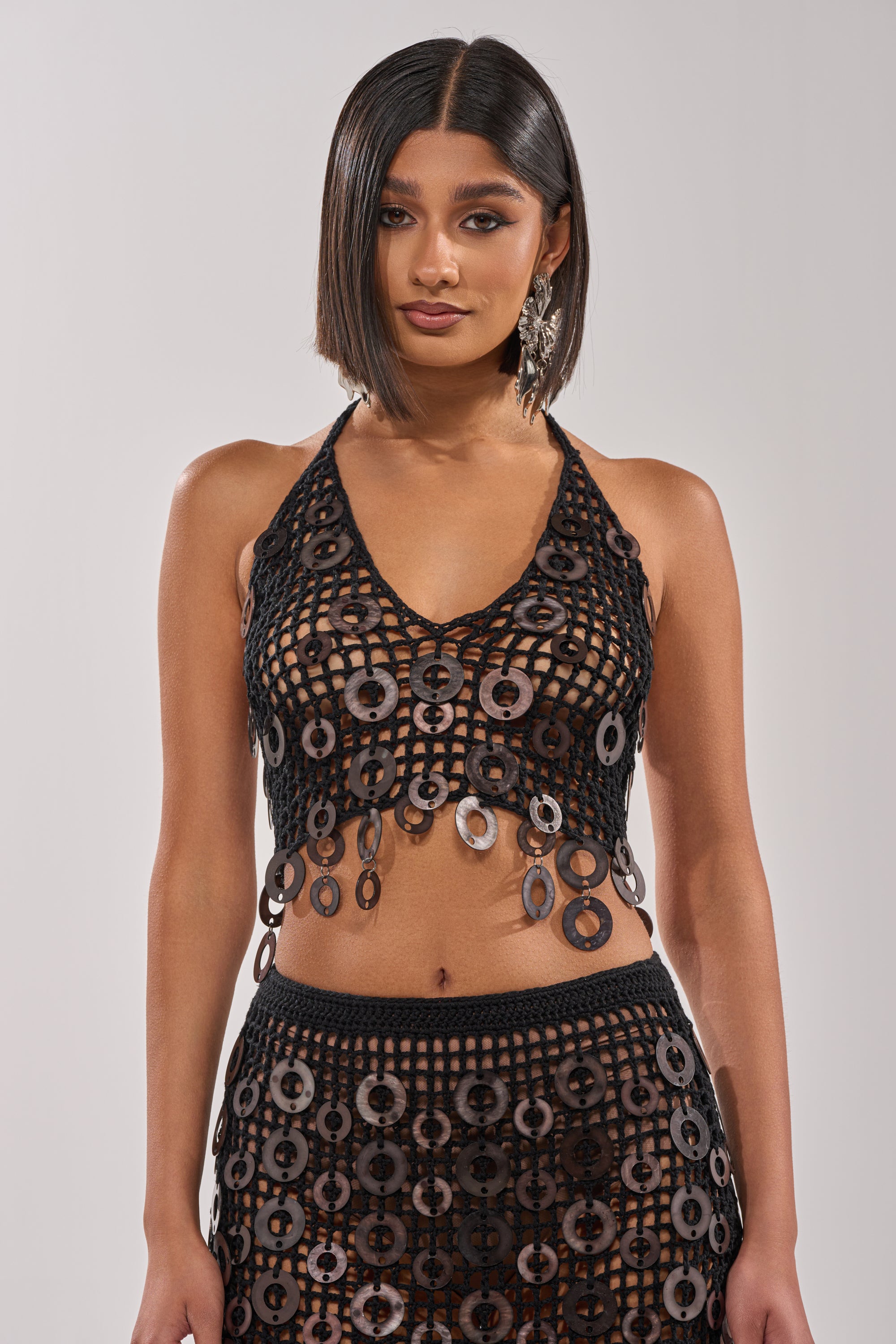 A woman with a sleek bob hairstyle wears the DESERT DANCER CROCHET HALTER TOP and matching skirt, both featuring large circular crochet patterns, making for an ideal festival look against a plain light gray background. 1