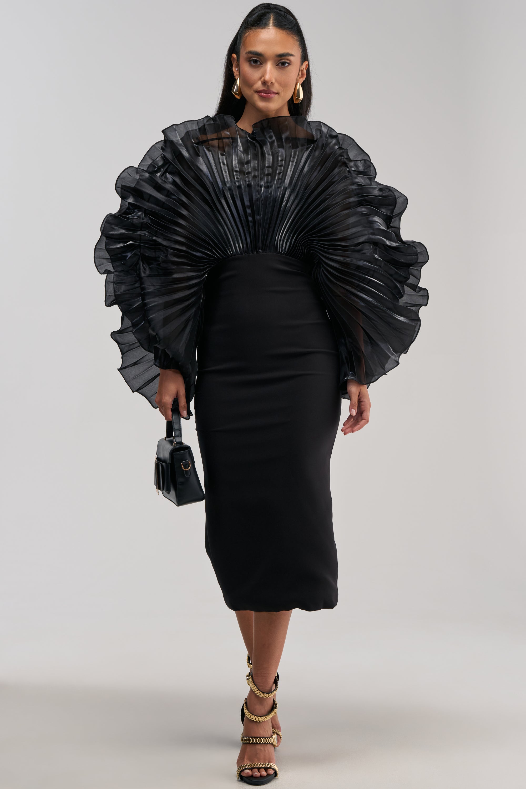 A woman wears the BY THE SEA SHORE BANDAGE MIDI DRESS IN BLACK, featuring a sculptural fan top and dramatic pleated organza ruffled sleeves, paired with gold hoop earrings, chain-detail heels, and a small black handbag, posed against a gray background.