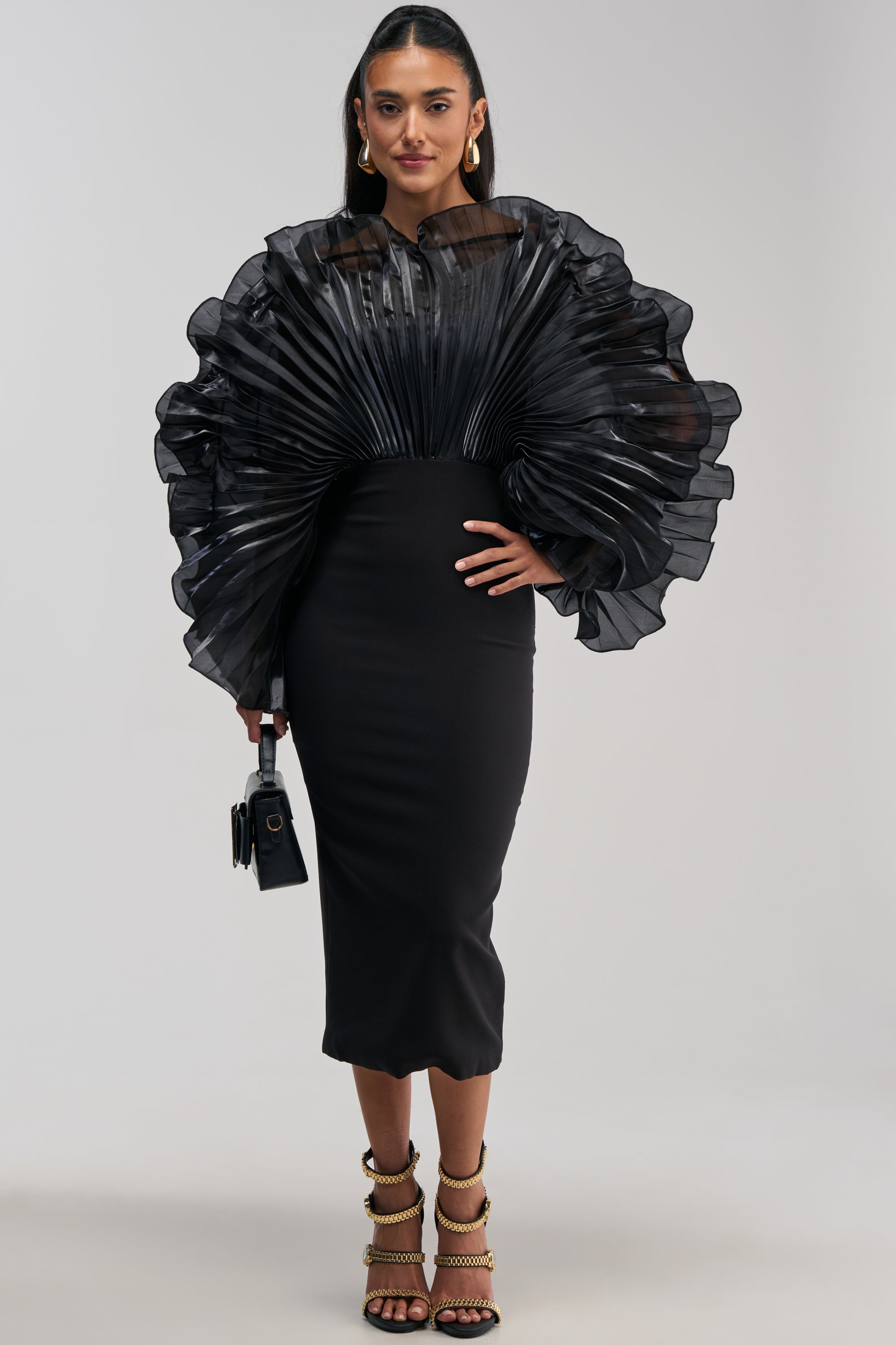 Woman in the BY THE SEA SHORE BANDAGE MIDI DRESS IN BLACK—a fitted dress with sculptural chiffon, dramatic pleated sheer fan-like sleeves—holding a small black handbag and wearing high-heeled gold-strapped sandals. She has long dark hair and hoop earrings.