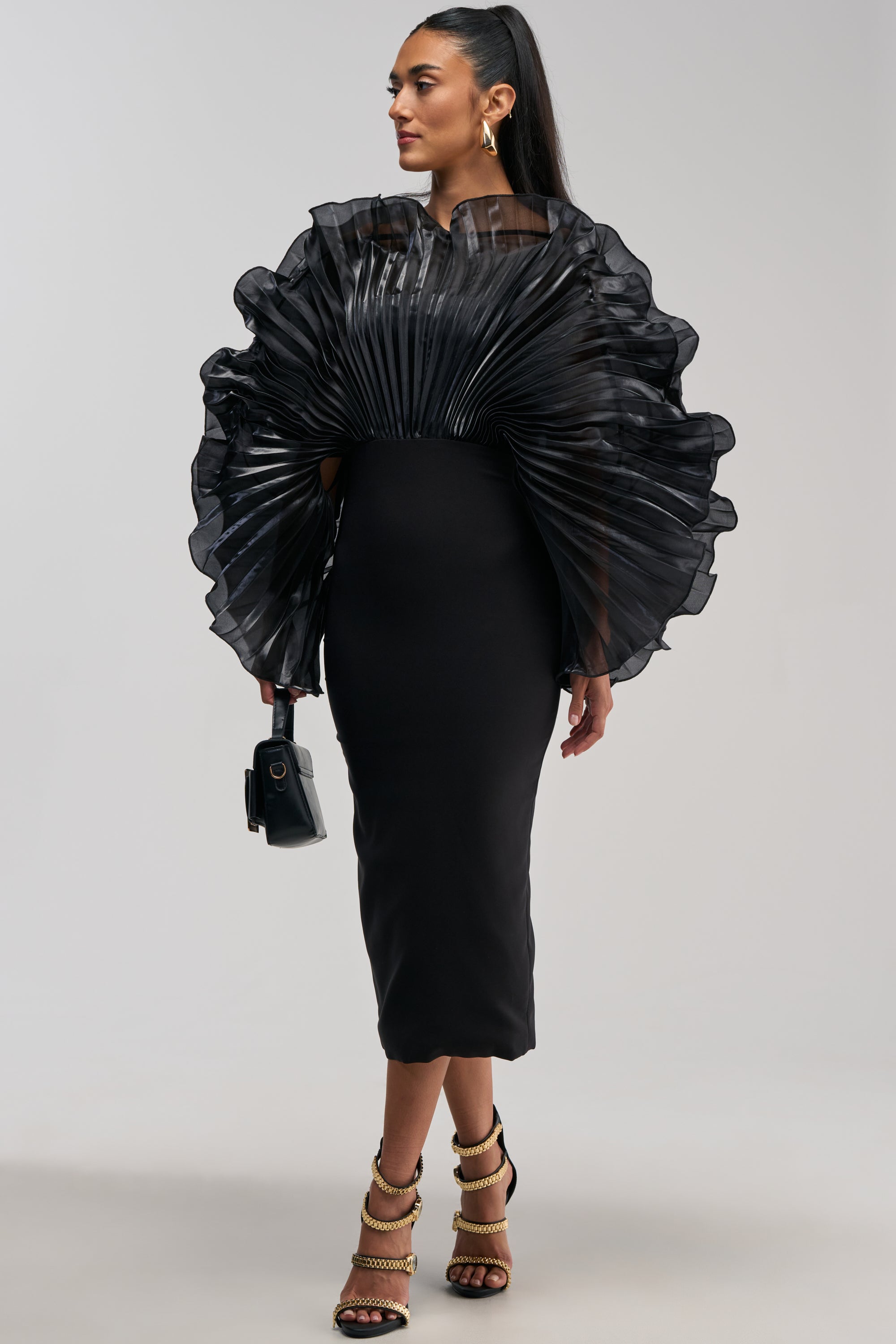 A woman stands against a plain light background wearing the BY THE SEA SHORE BANDAGE MIDI DRESS IN BLACK, a fitted midi dress featuring dramatic pleated sheer ruffles at the shoulders and chest. She pairs the look with a black handbag and gold strappy heels.