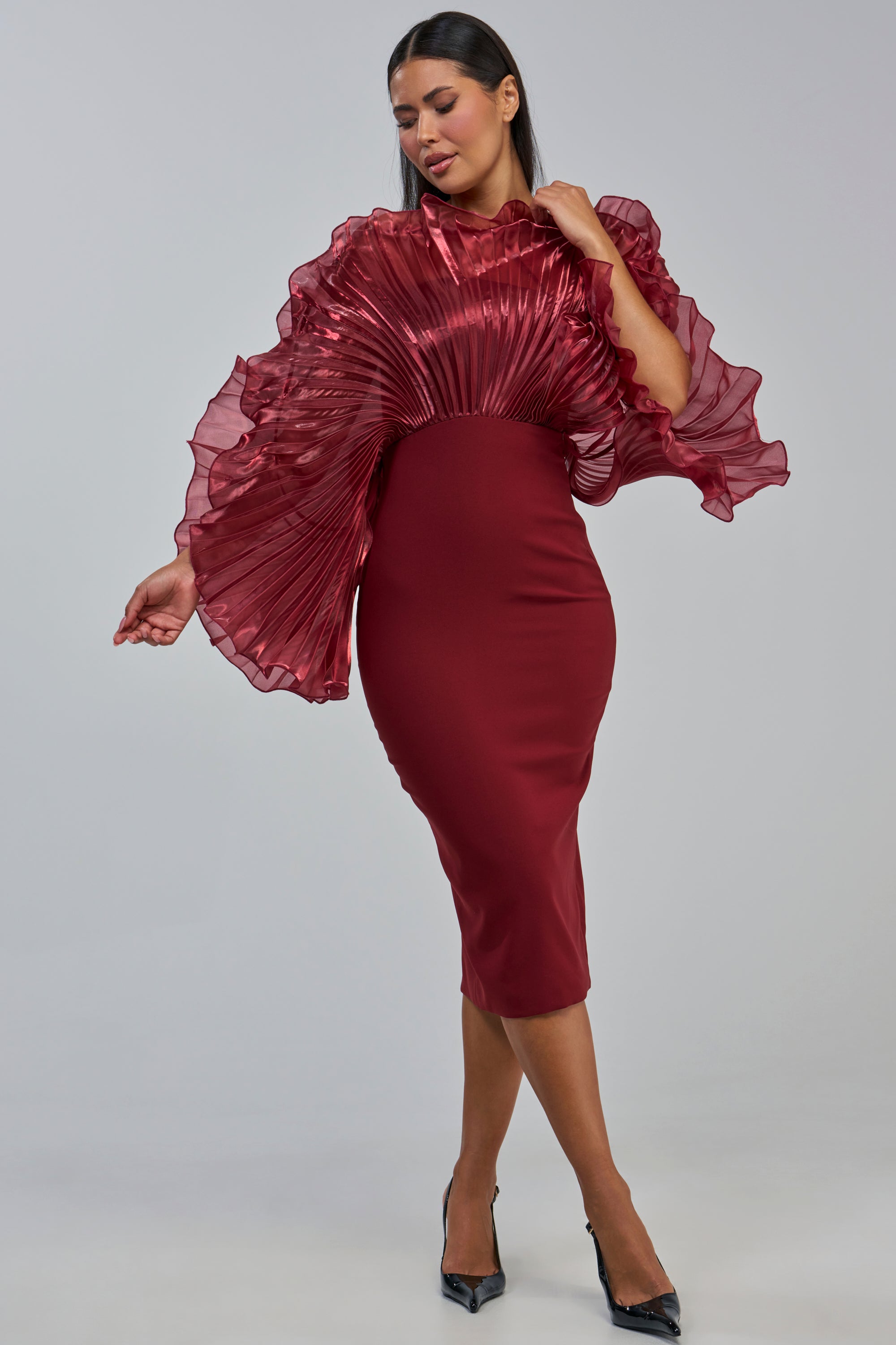 A woman stands against a plain light gray background, wearing the BY THE SEA SHORE Bandage Midi Dress in Burgundy, featuring a fitted silhouette and dramatic pleated organza ruffles defining the shoulders and sleeves.