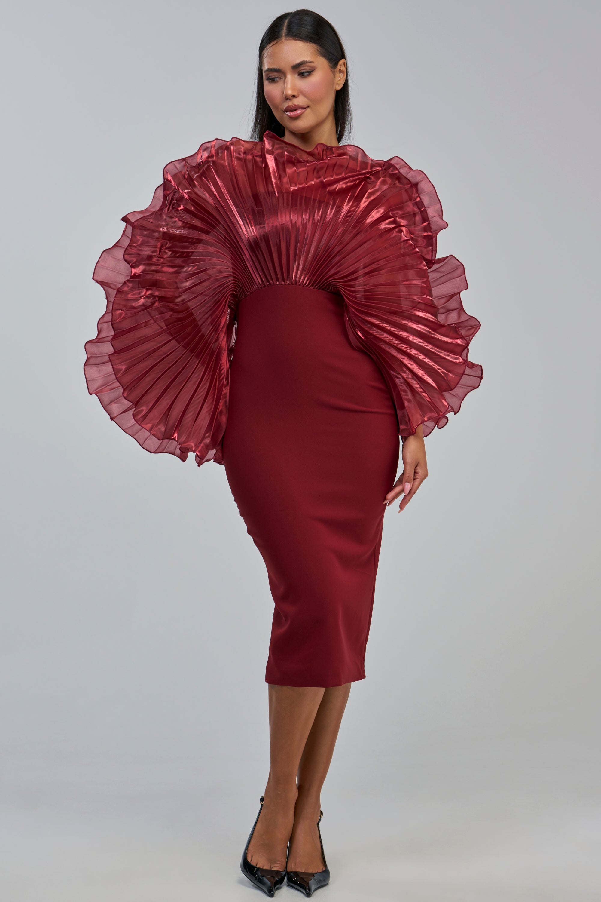 A woman wearing the BY THE SEA SHORE BANDAGE MIDI DRESS IN BURGUNDY, featuring a fitted silhouette and dramatic pleated organza ruffles on the shoulders and upper bodice, poses against a plain light gray background.
