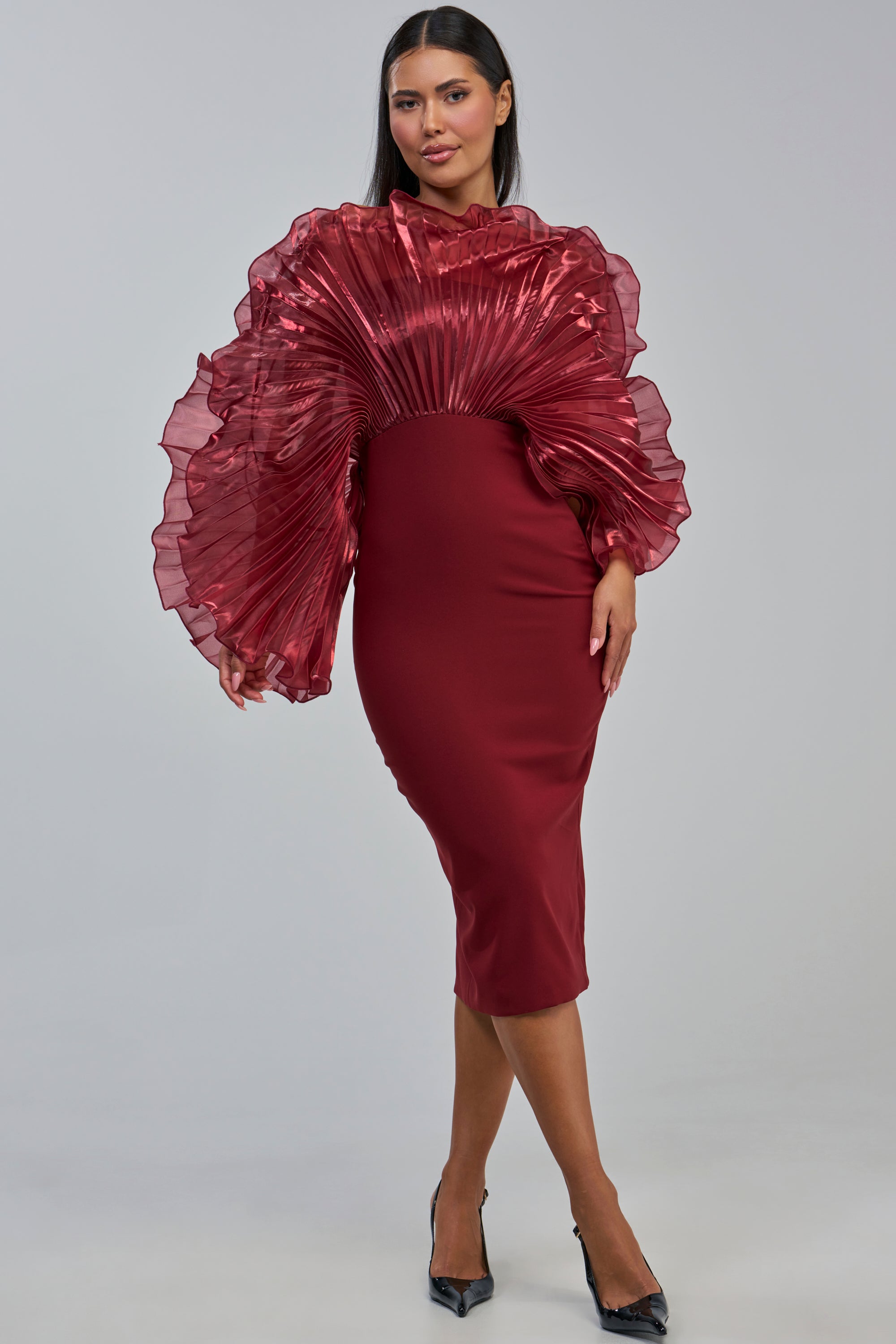 A woman poses confidently against a plain light gray background in the BY THE SEA SHORE BANDAGE MIDI DRESS IN BURGUNDY, a fitted, knee-length dress featuring dramatic pleated ruffled shiny sleeves.