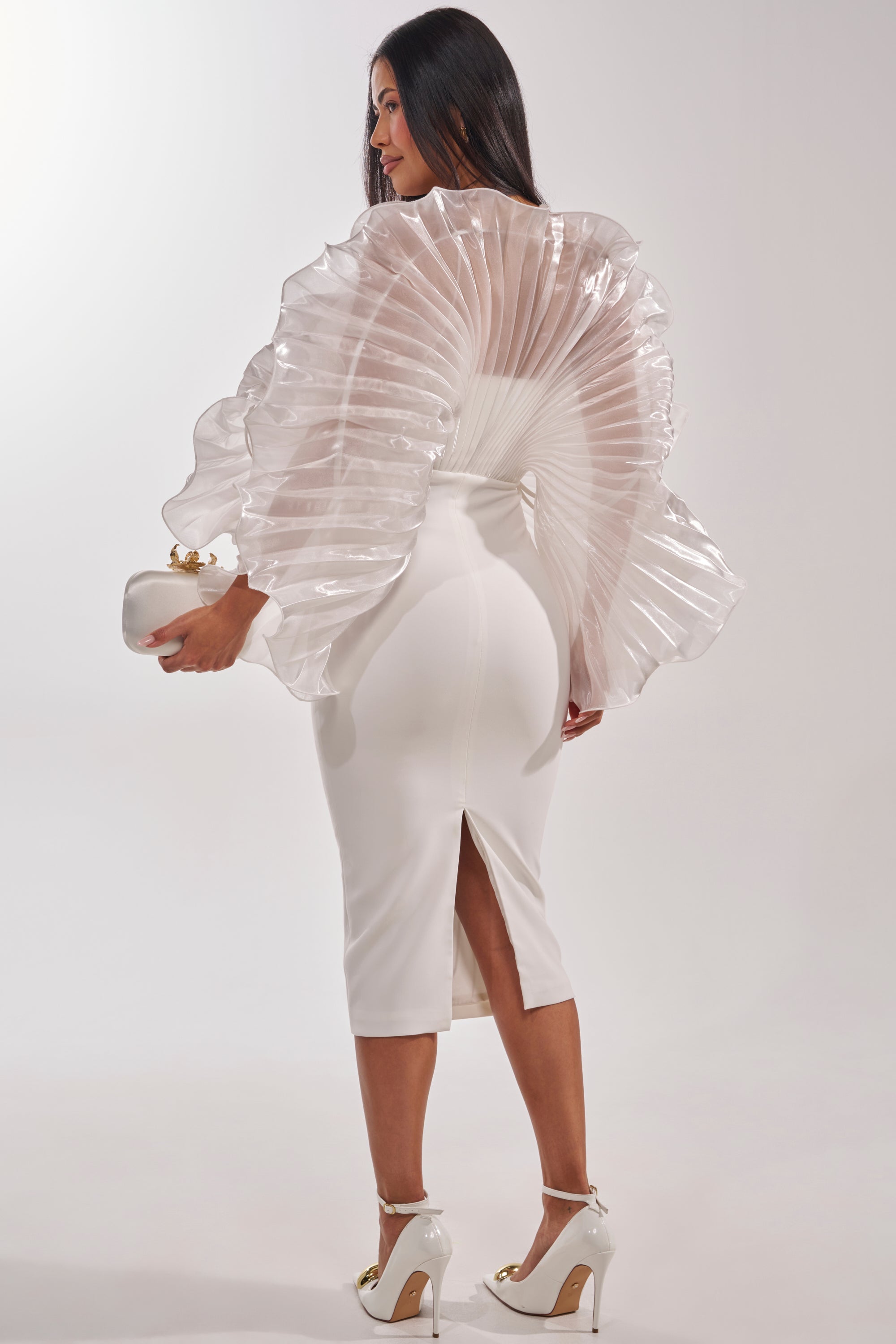 A woman in the BY THE SEA SHORE BANDAGE MIDI DRESS with a dramatic ruffled cape holds a white clutch and wears white high heels with gold accents, standing against a plain background. 5
