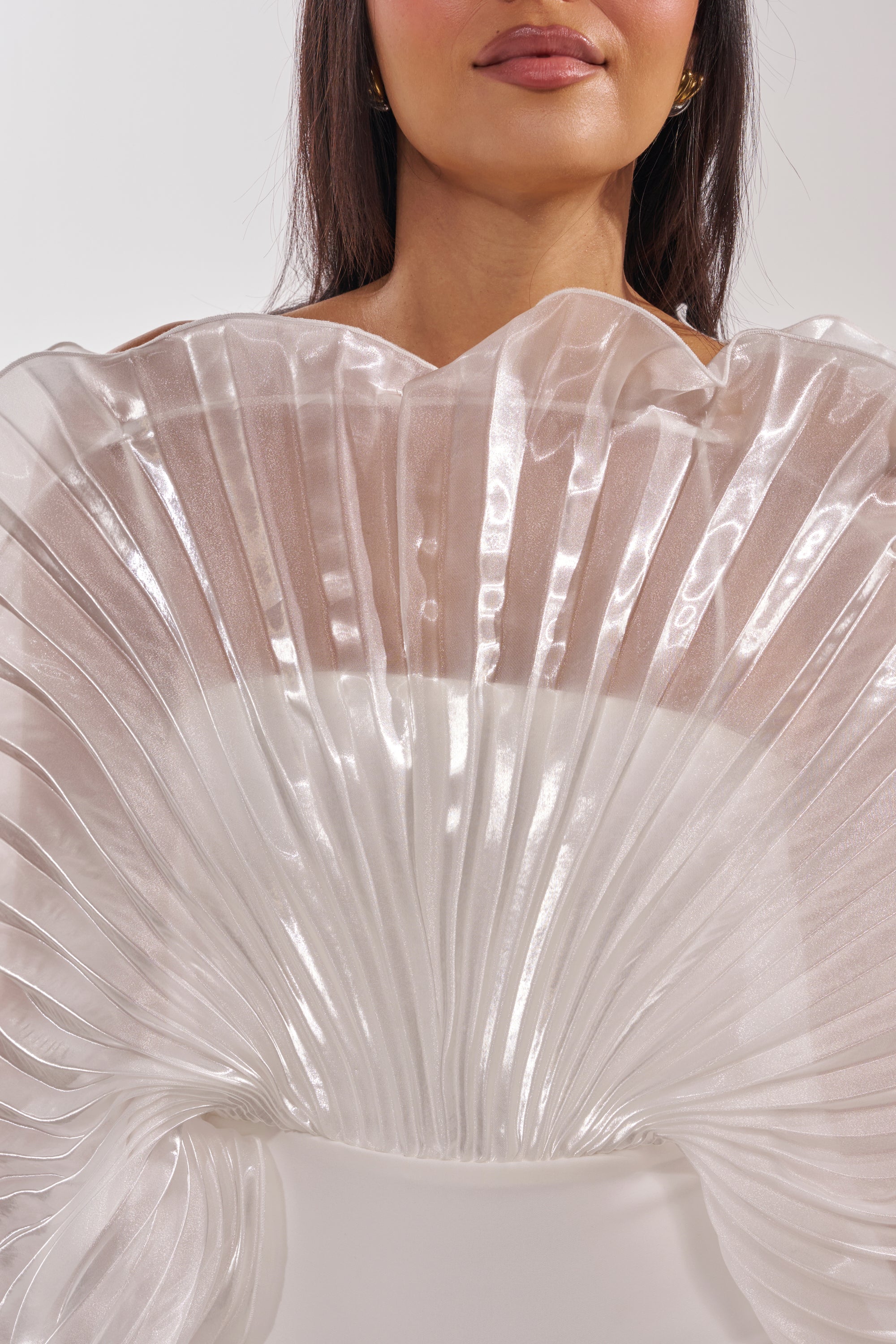 A woman wears the BY THE SEA SHORE BANDAGE MIDI DRESS—a structured, fan-pleated, translucent white dress with a glossy sculptural design around the shoulders and chest—shown from the shoulders up against a light background. 6