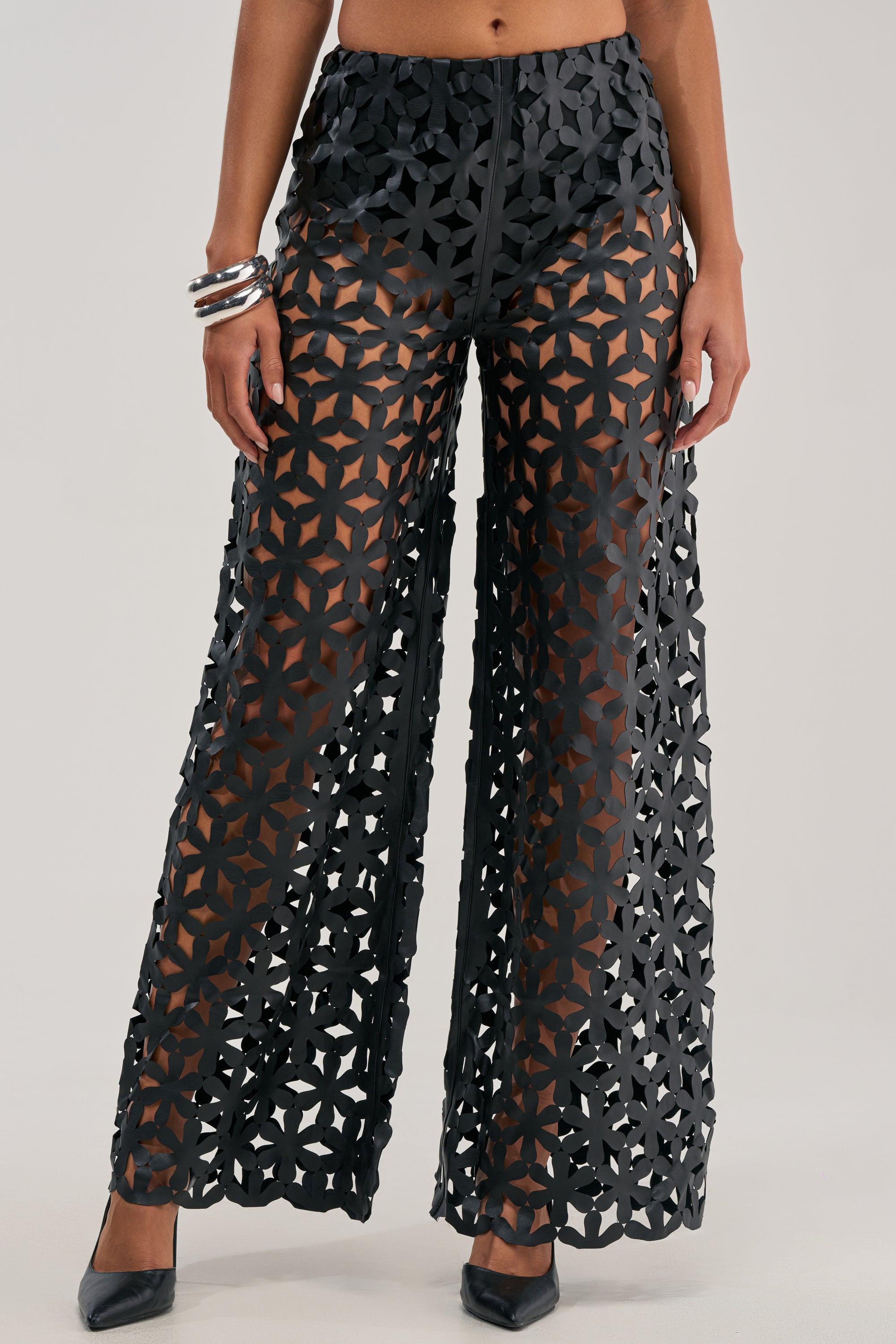 A woman models the LEAH TROUSER IN BLACK, featuring a geometric lattice cutout design that reveals her legs underneath. She accessorizes with silver bangles on one arm and black pointed-toe heels. Only her torso and legs are visible.