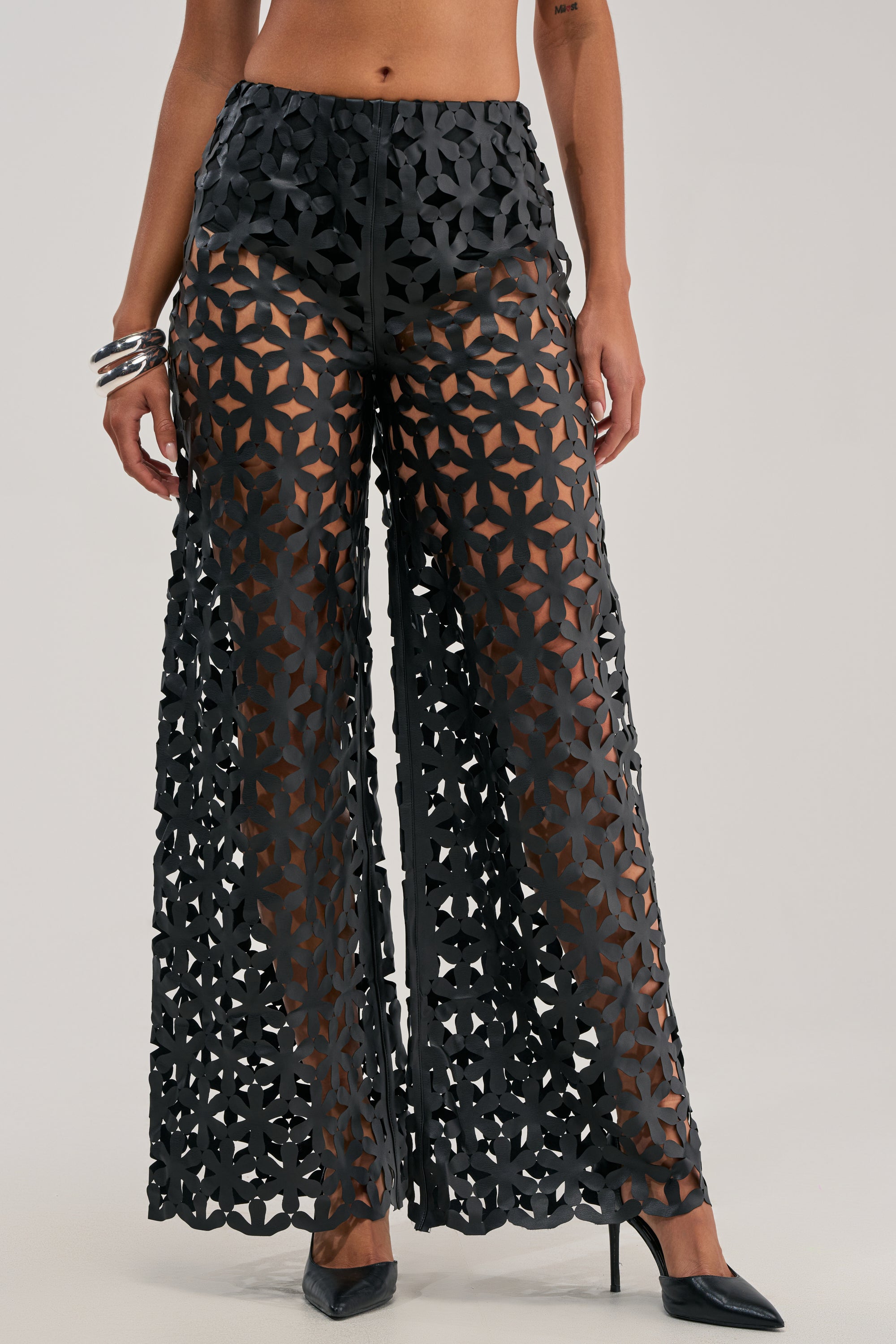 A person wearing the LEAH TROUSER IN BLACK—high-waisted, black, sheer wide-leg lattice cutout pants with a geometric pattern. The top portion of these trousers is lined. The outfit is styled with black high heels and silver bangles, set against a plain light background.