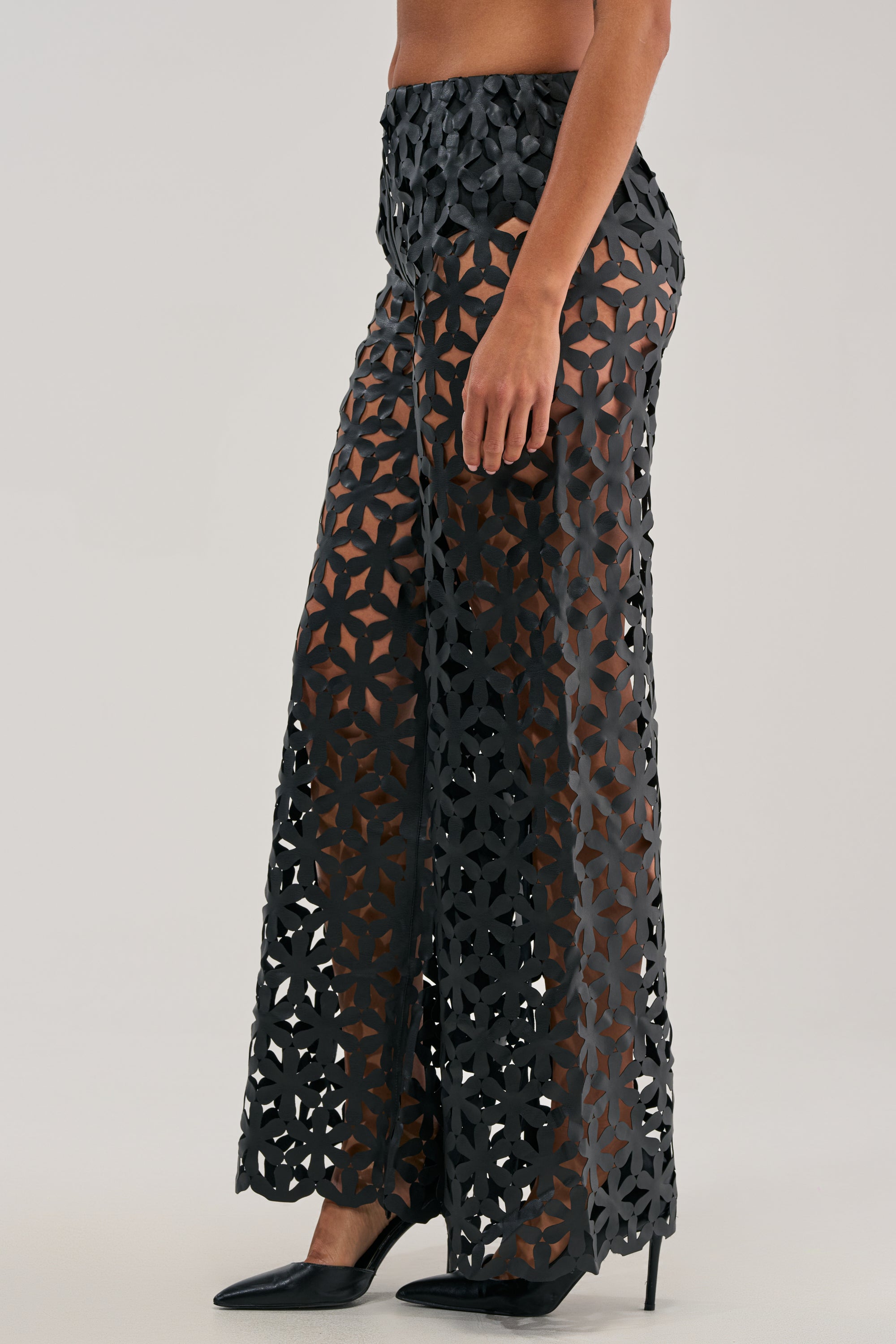 A person wearing the LEAH TROUSER IN BLACK, featuring a high-waisted fit with geometric lattice cutouts, paired with black pointed-toe heels and standing against a plain background.