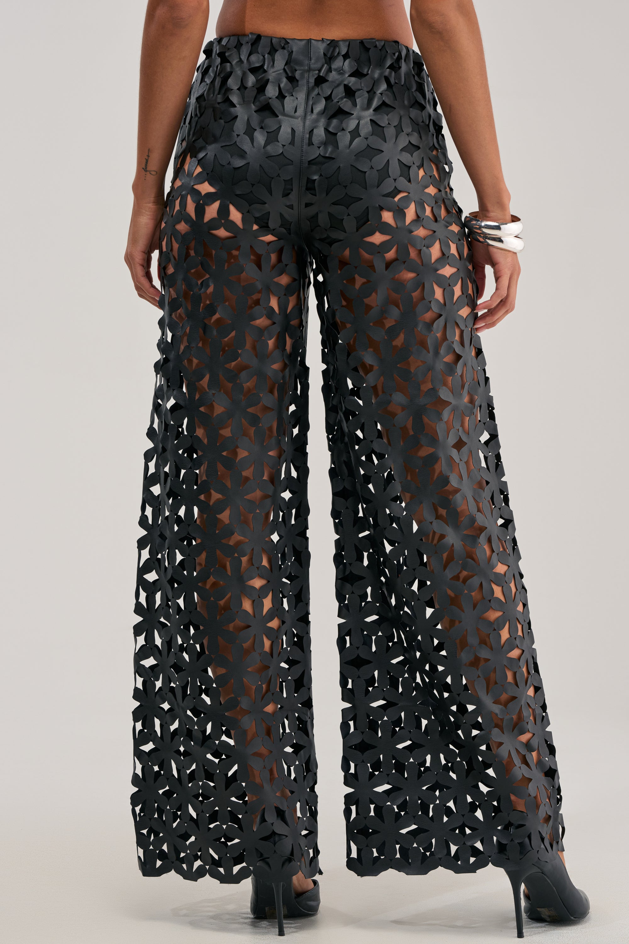 Wearing the LEAH TROUSER IN BLACK—high-waisted pants featuring a geometric lattice cutout design that reveals skin beneath—paired with high heels and a bracelet. The photo is captured from behind.