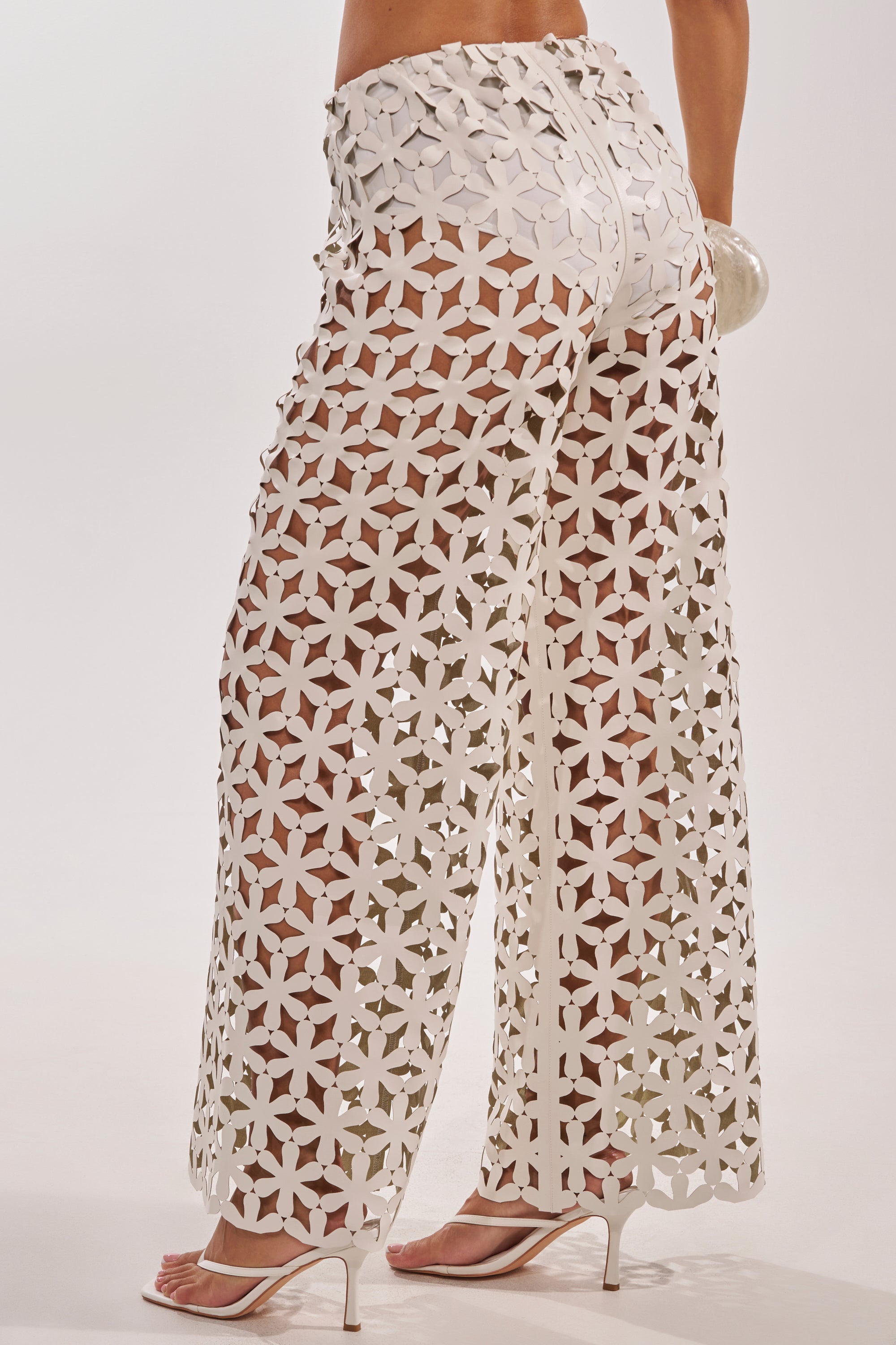 Wearing the LEAH TROUSER—high-waisted, wide-leg white pants with floral cutout lace that reveal white underwear—the person pairs them with white heeled sandals and carries a small round purse. 3