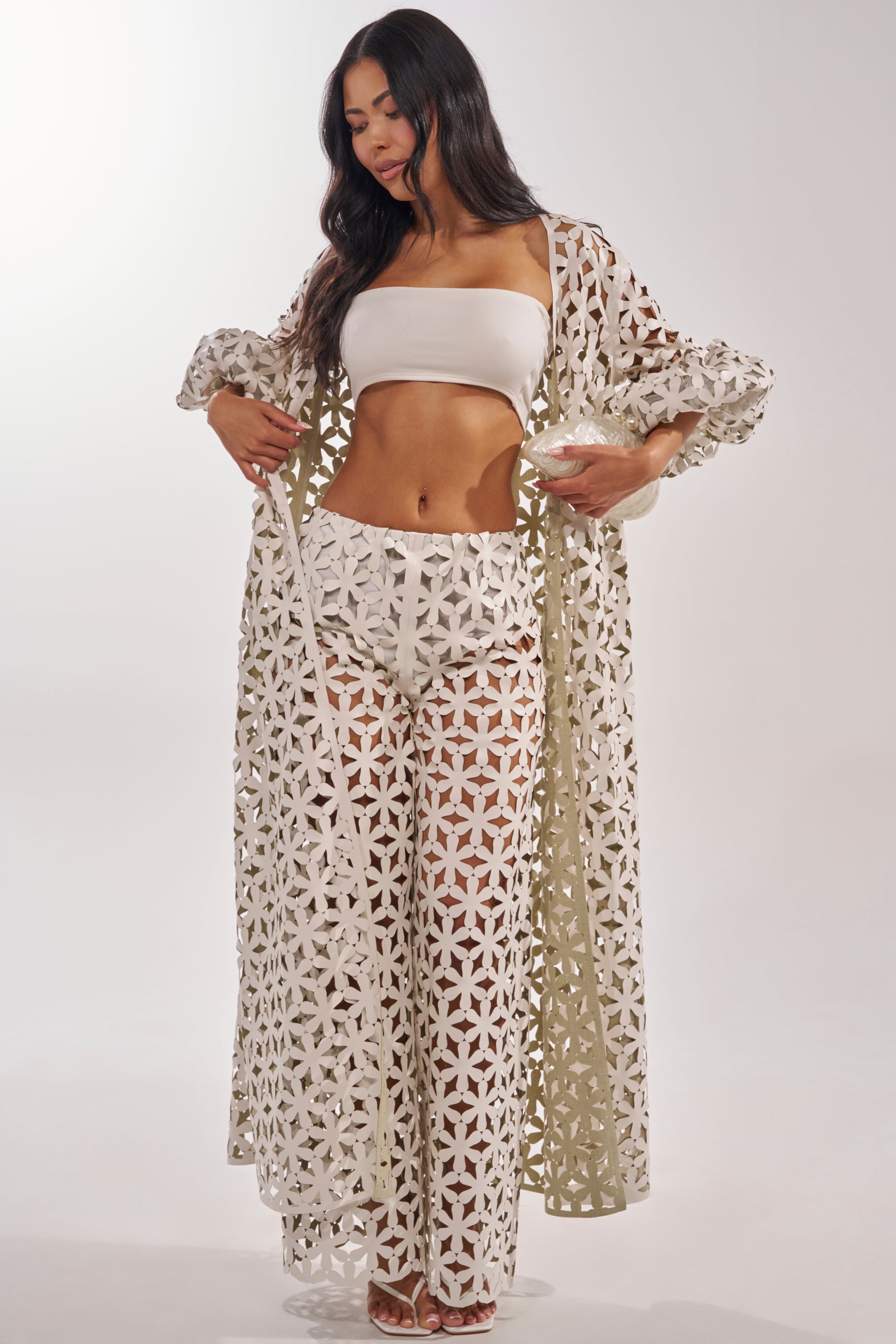 Woman wearing LEAH TROUSER paired with a white bandeau top and long, sheer, floral lace cover-up; standing against a plain light background and looking downwards. 6
