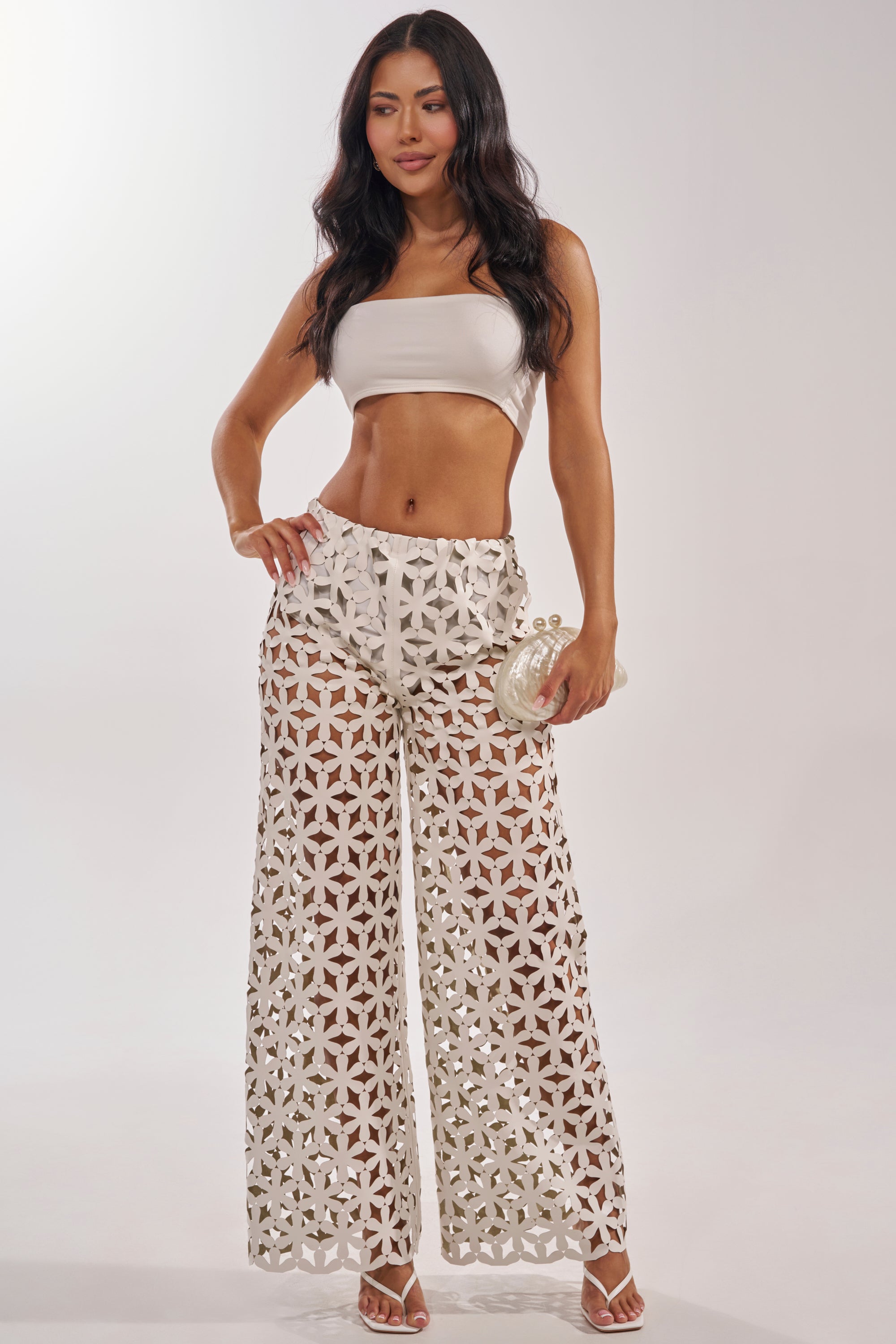 A woman poses in the LEAH TROUSER, featuring a geometric pattern and high-waisted wide-leg style. She pairs it with a white bandeau top, white heeled sandals, and carries a small clutch against a plain light background. 7