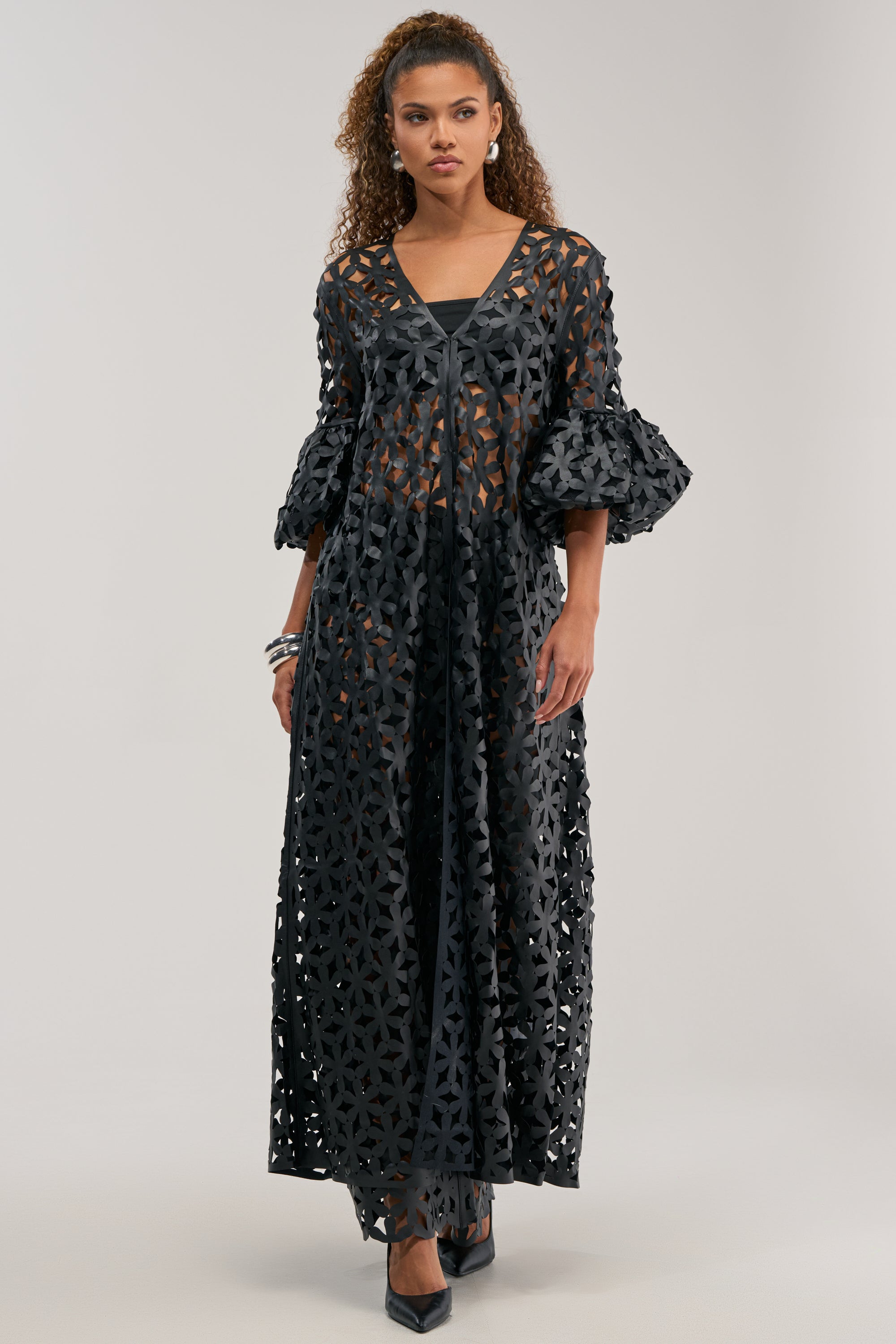 A woman stands against a plain light background, layering the LEAH DUSTER IN BLACK—a longline statement piece—over a long, sheer black dress with geometric cutouts and voluminous three-quarter sleeves.