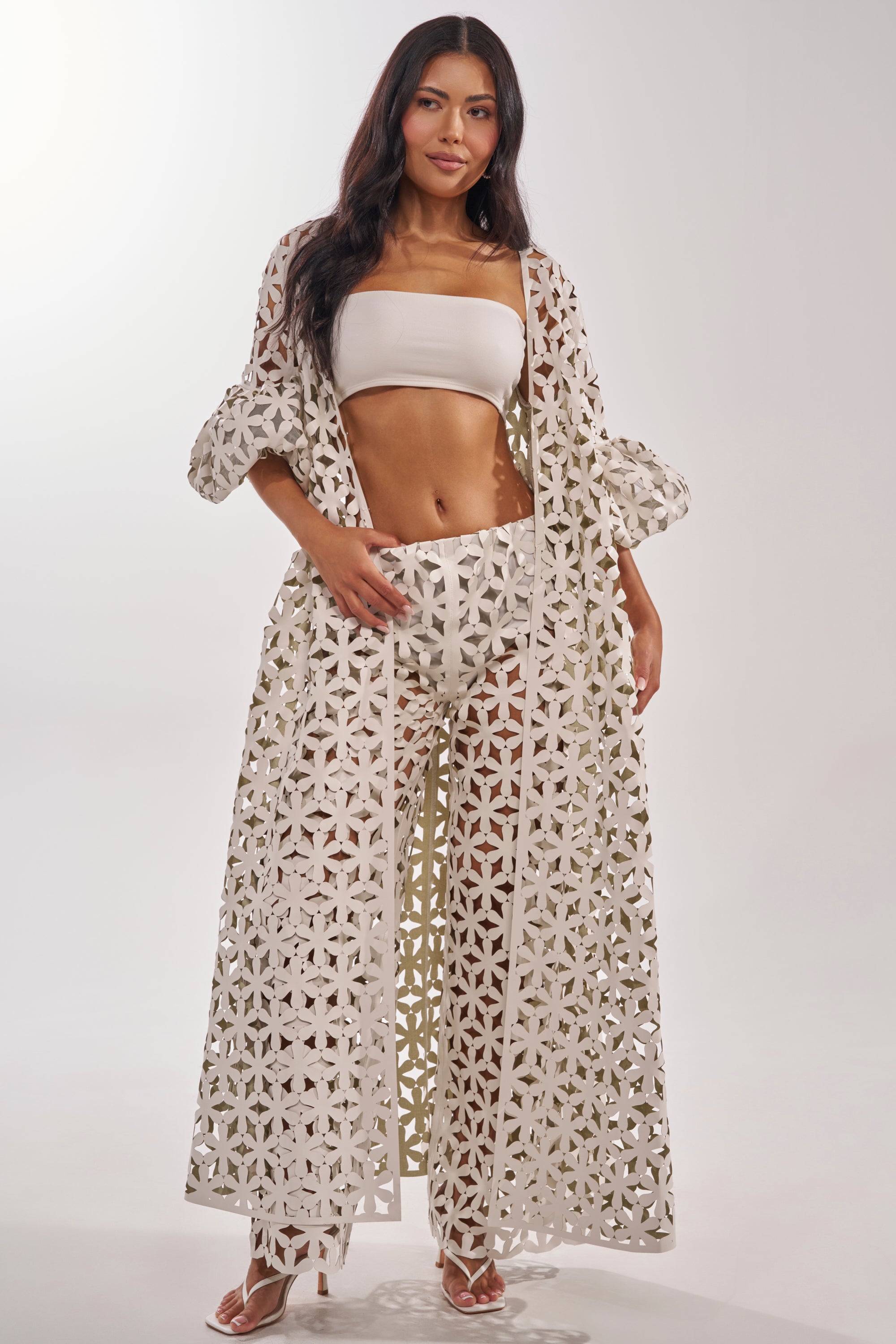 A woman with long, dark hair poses confidently in the LEAH DUSTER—a long, white faux leather duster with a lattice pattern—styled over a bandeau top and wide-leg pants against a plain light background. 1