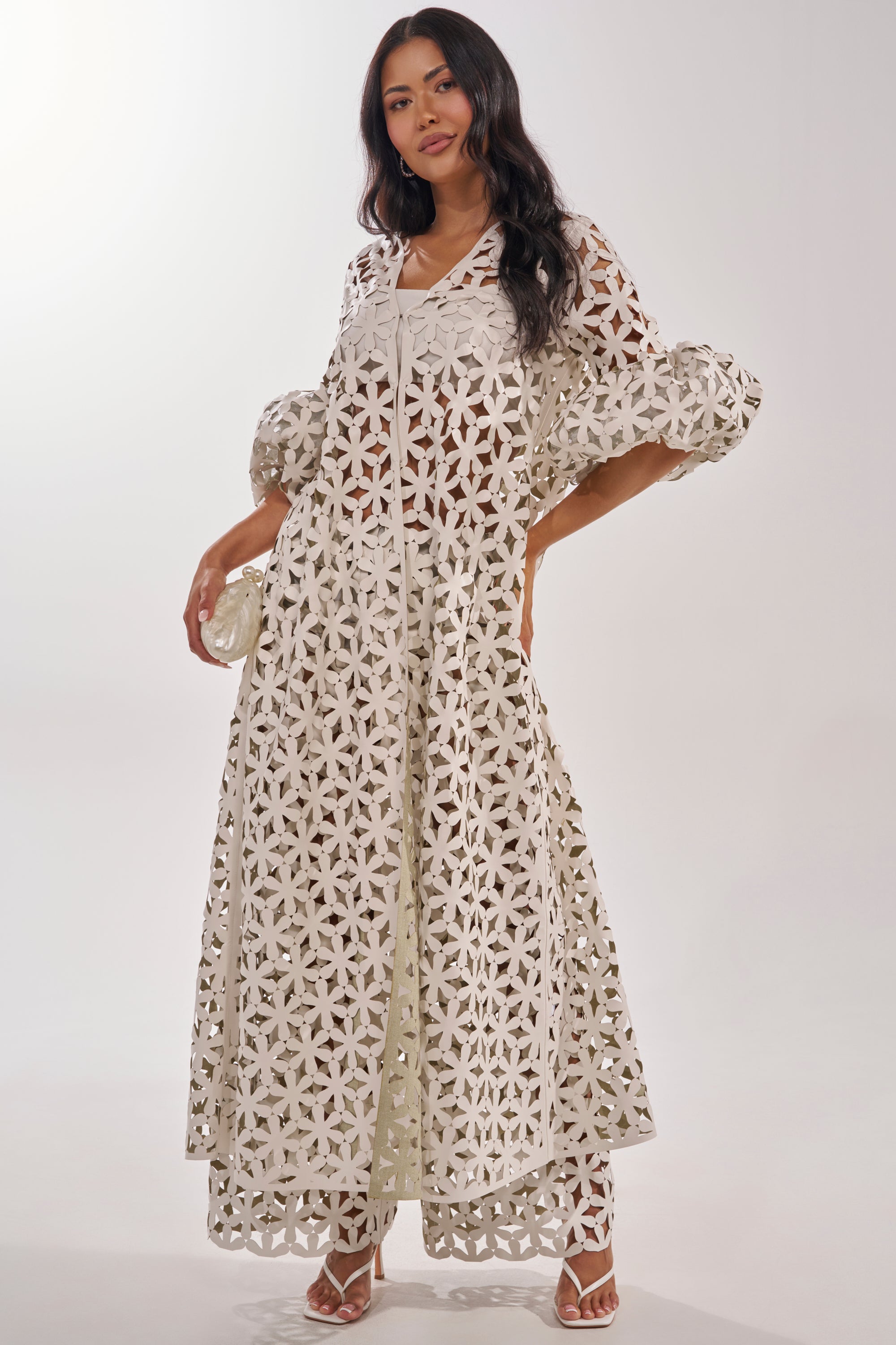 A woman poses in the LEAH DUSTER, a long white dress with geometric cutouts and puffed sleeves, styled with white heels and a small chic lattice handbag, all set against a plain light background. 3