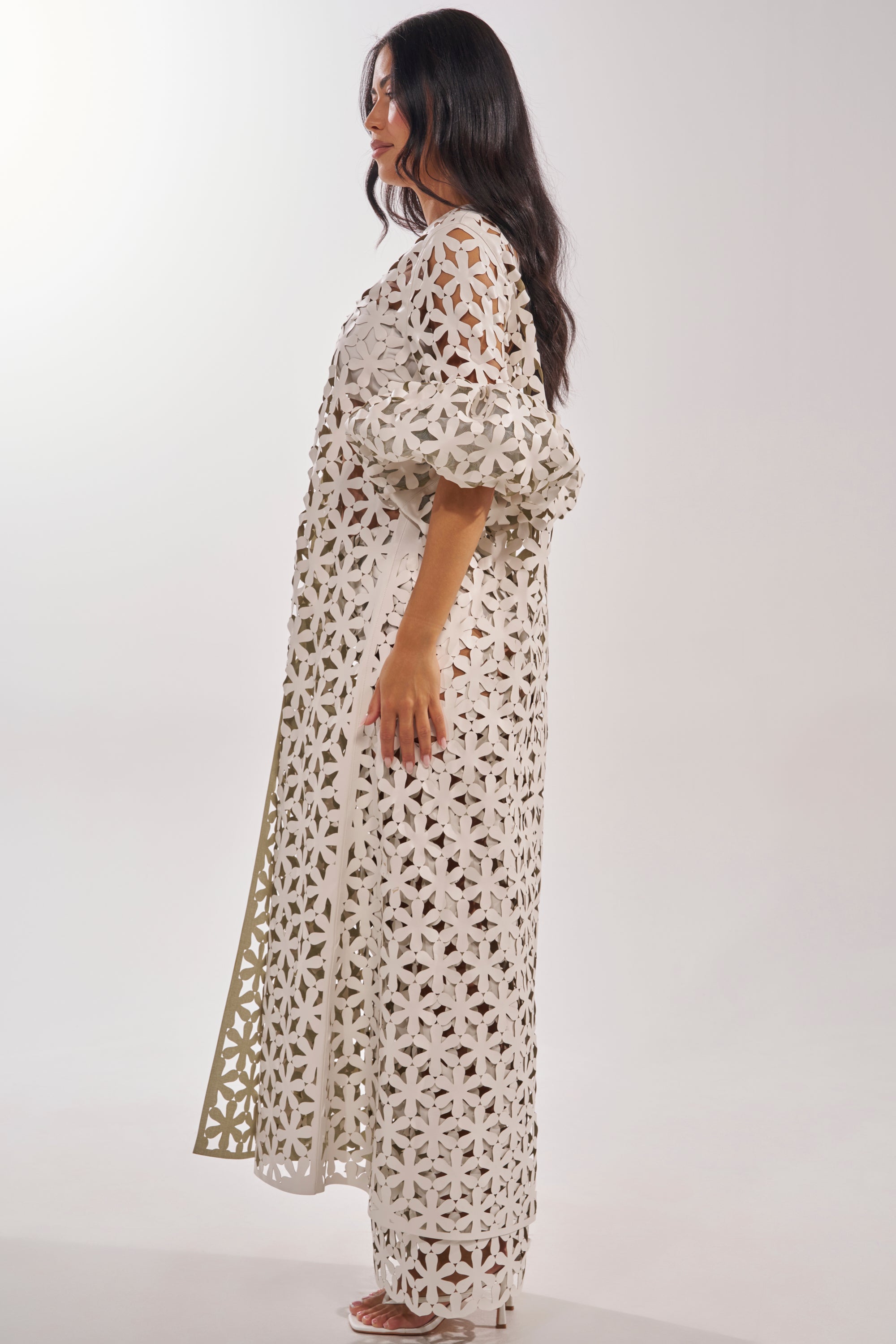 A woman with long dark hair stands in profile against a plain white background, wearing the LEAH DUSTER—resort wear featuring a loose-fitting, cream-colored design with an intricate geometric cut-out pattern. 4