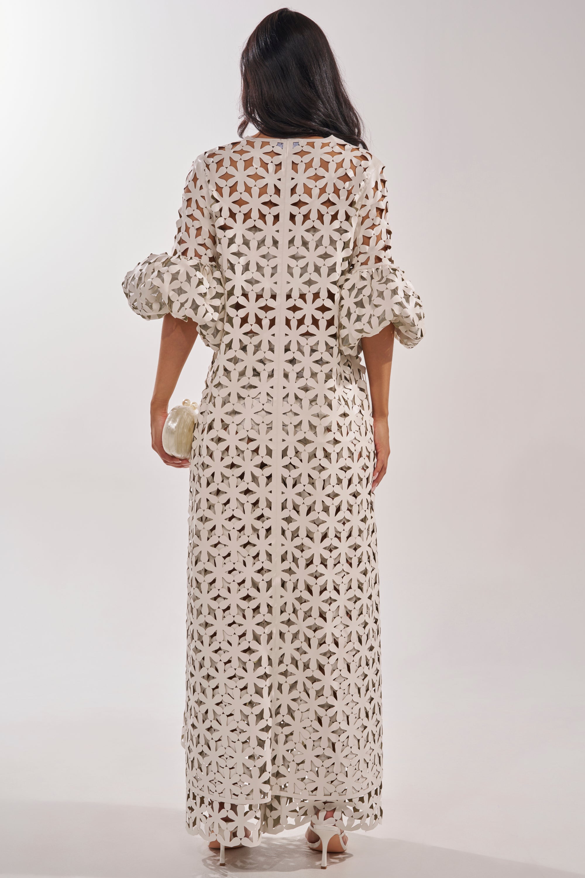 A woman with long dark hair stands facing away, wearing the LEAH DUSTER—a long off-white lace resort dress with a geometric floral pattern and puffed sleeves. She pairs it with white heels and holds a small white clutch against a plain light background. 5