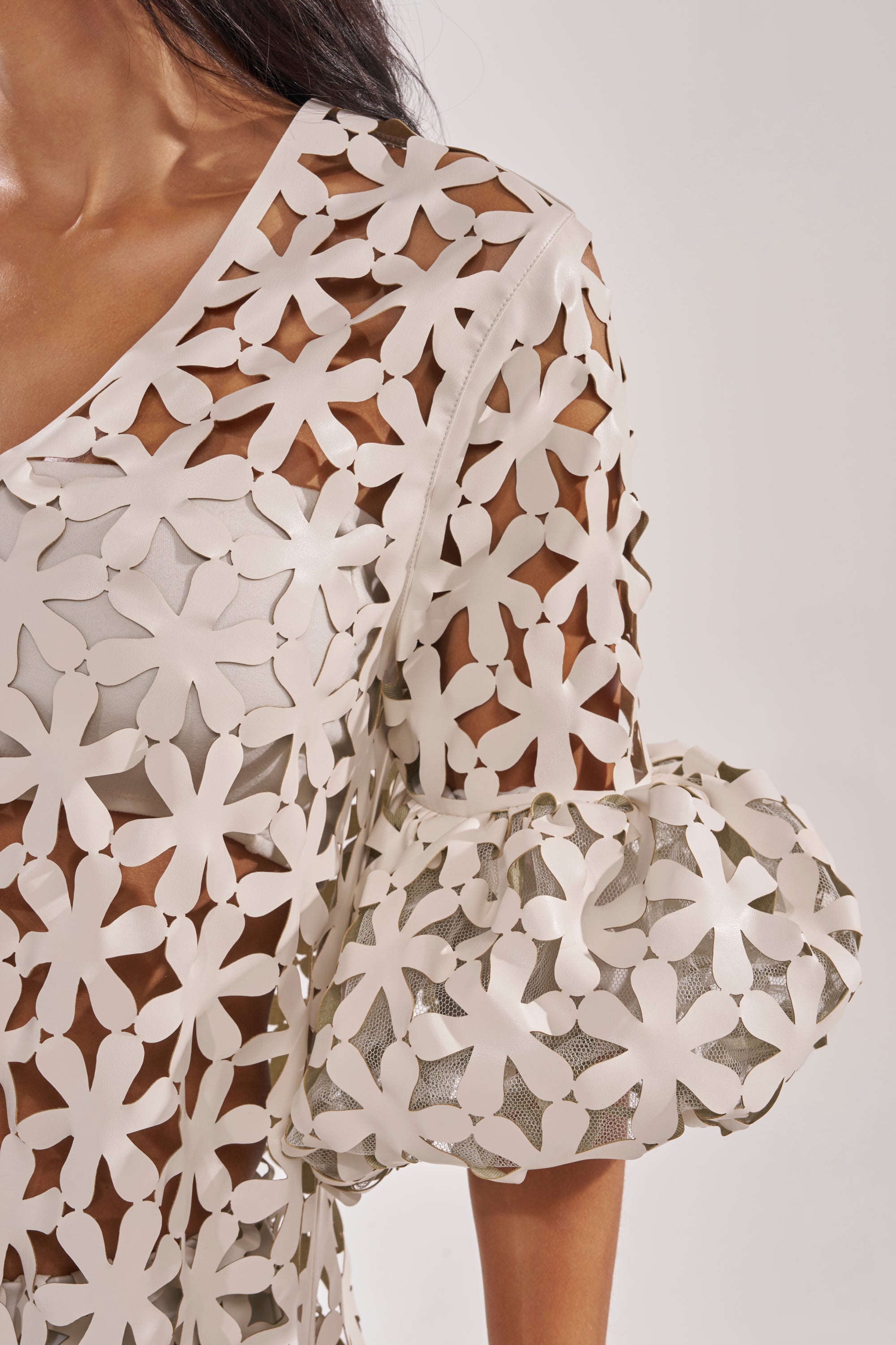 A close-up of a woman in resort wear: she wears the LEAH DUSTER, a cream-colored top with a white lattice pattern and puffed sleeves, revealing glimpses of bare skin beneath. 6