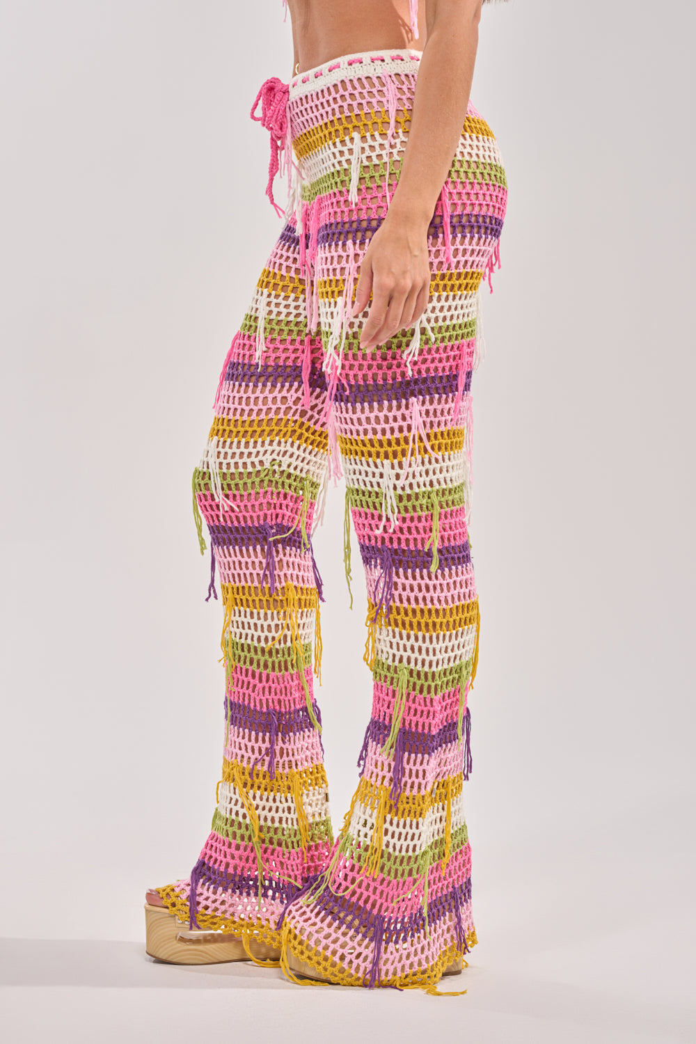 A person models the RESORT BABE CROCHET PANT, featuring vibrant pink, yellow, green, and white stripes, paired with platform sandals against a plain light background. 2