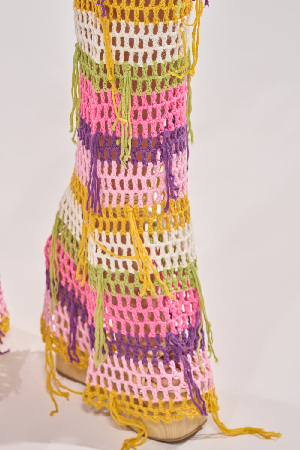 Close-up of RESORT BABE CROCHET PANT, featuring flared legs with pink, purple, yellow, white, and green stripes plus loose fringe accents—styled over tan platform shoes for chic beachwear appeal. 6