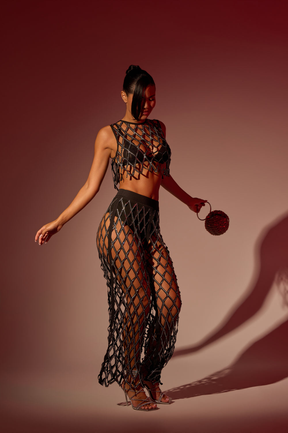A woman poses under red lighting in the ROCK N ROLL RHINESTONE TROUSER and a matching mesh crop top over a black bikini, accessorized with a small round bag and strappy heels. 1