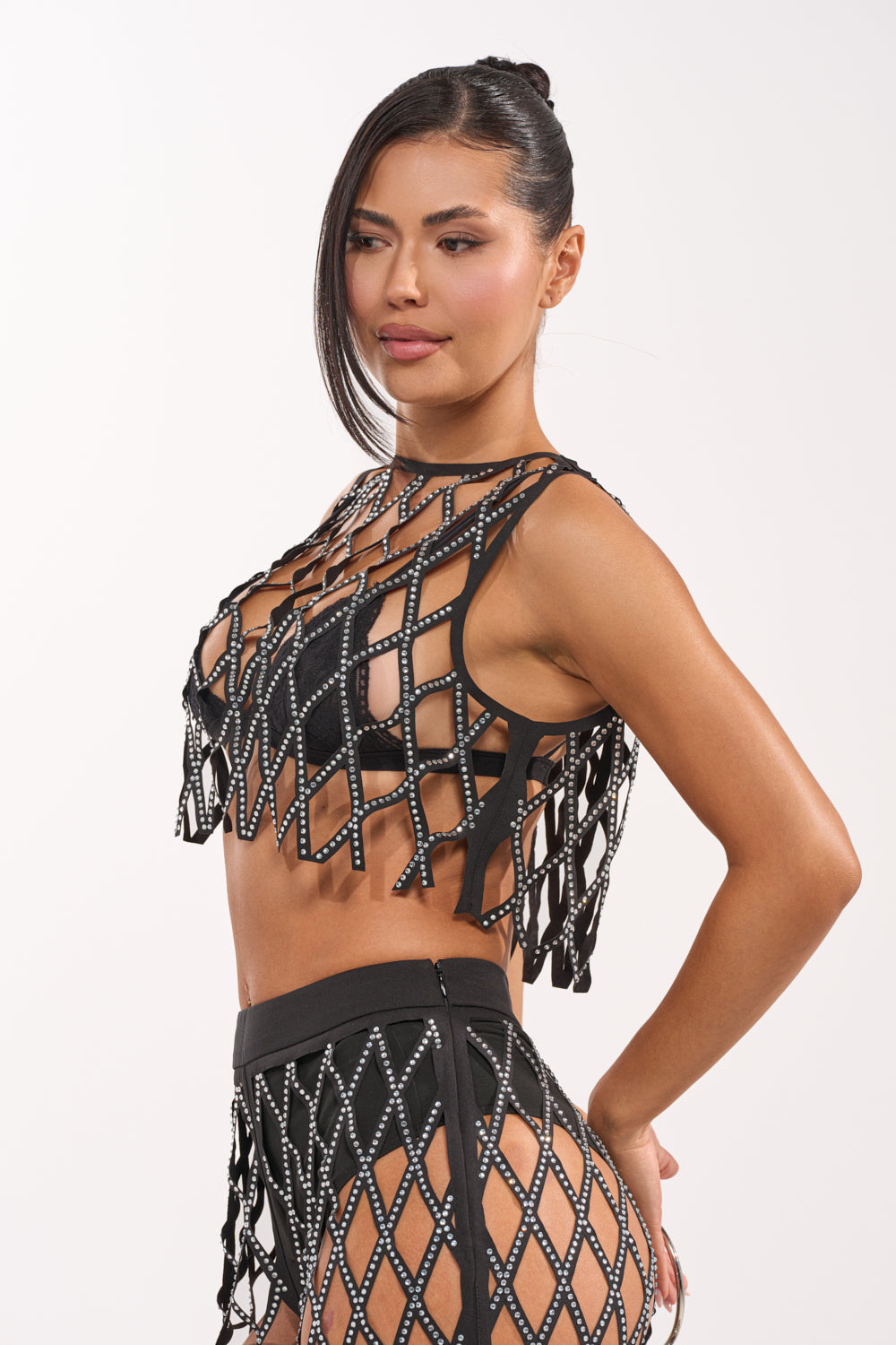 A woman models the ROCK N ROLL RHINESTONE TOP, a black fishnet-style crop top with large diamond-shaped openings, paired with high-waisted bottoms. Her long dark hair is pulled back as she poses against a plain white background. 4