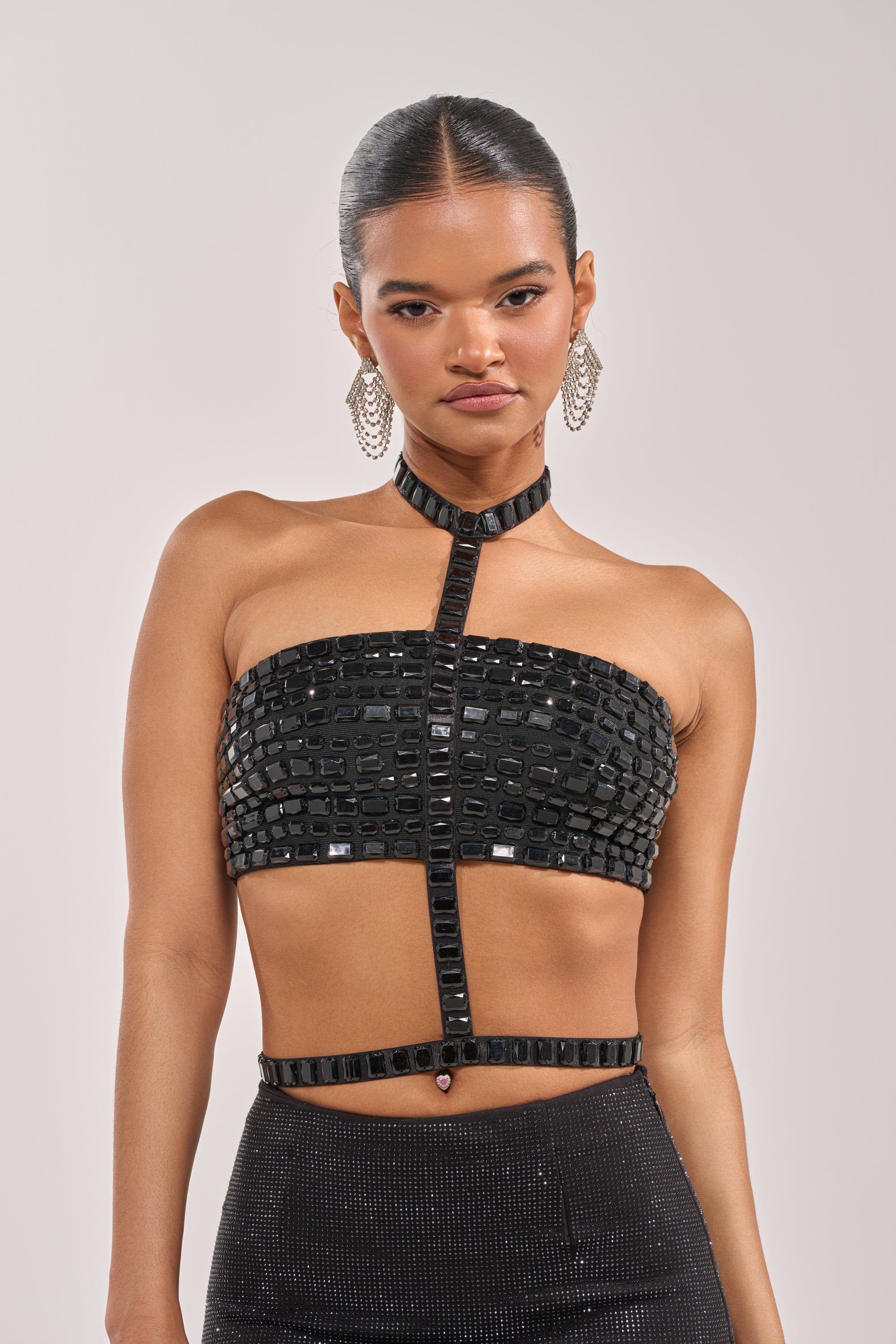 A woman with slicked-back hair models the OH HOT DAMN BRALETTE, featuring a strapless embellished design, vertical choker detail, dangling earrings, and a high-waisted black skirt against a plain background. 0