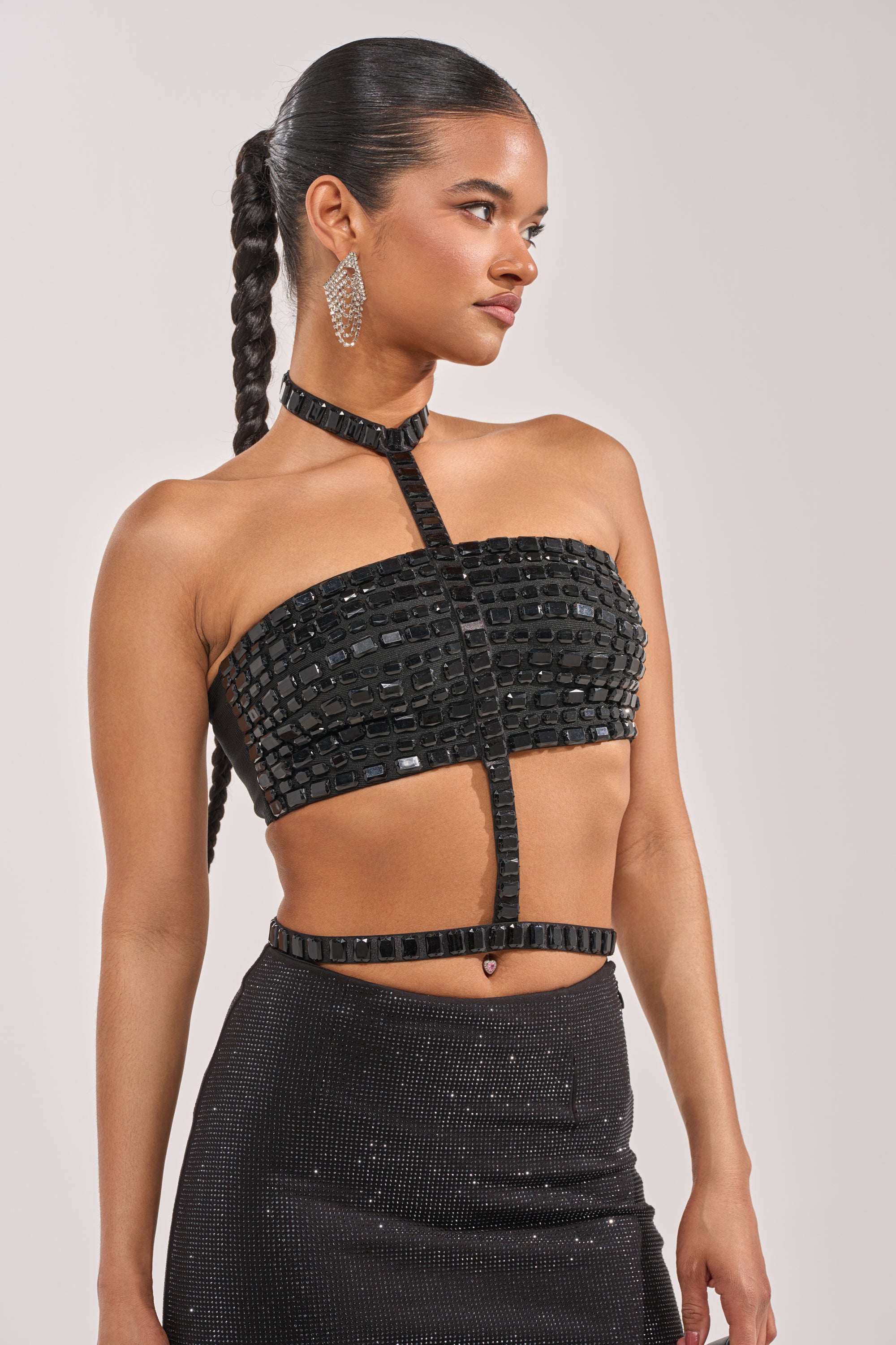 A woman with long, braided hair wears the OH HOT DAMN BRALETTE—a black, strapless crop top with square sequins and a matching waistband. Sparkly earrings and a high-waisted, shimmering skirt with black gemstones complete her confident look. 1