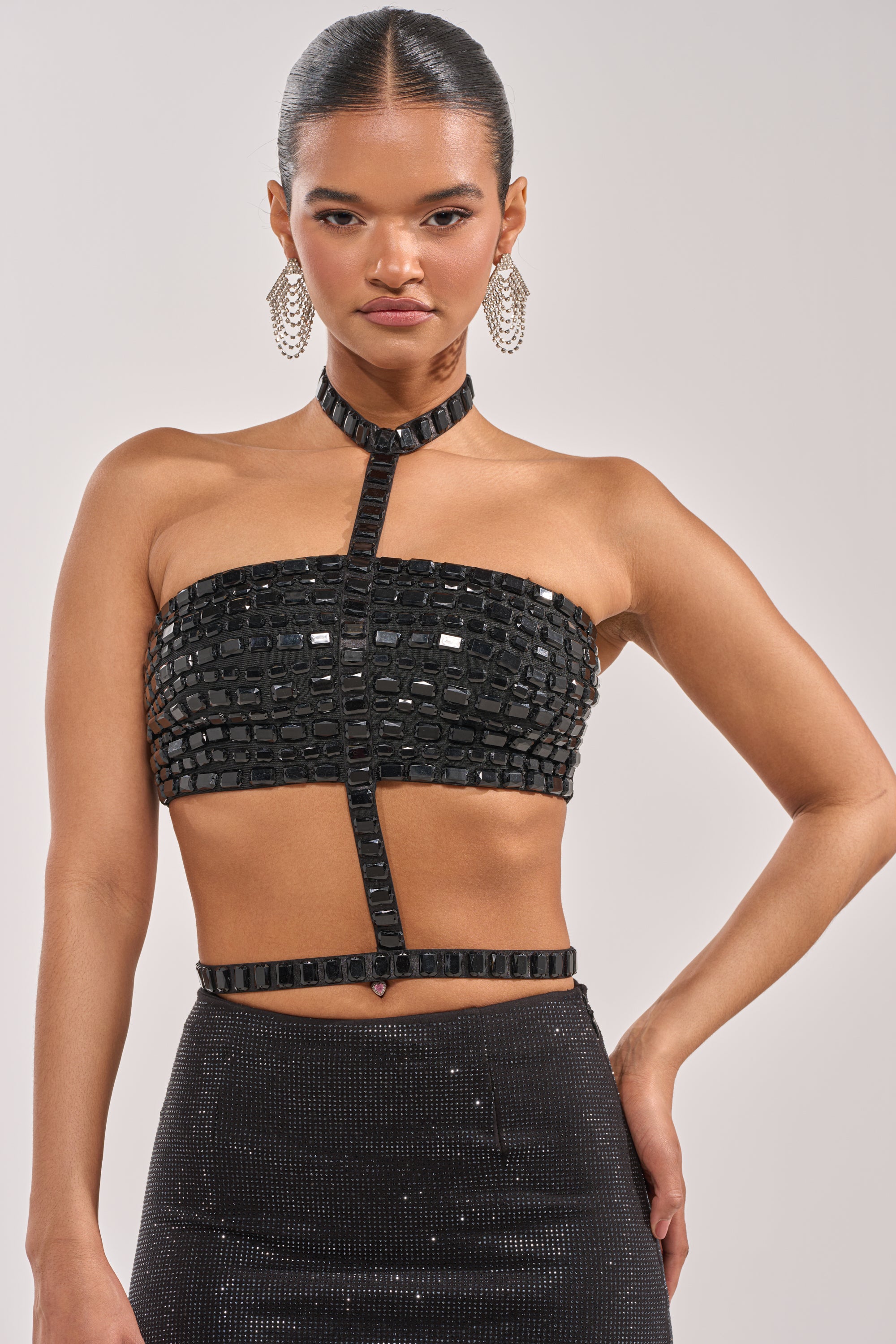 A woman with slicked-back hair wears the OH HOT DAMN BRALETTE, a strapless party top with shiny square embellishments and a choker with vertical strap, paired boldly with large earrings and a high-waisted black skirt. 2