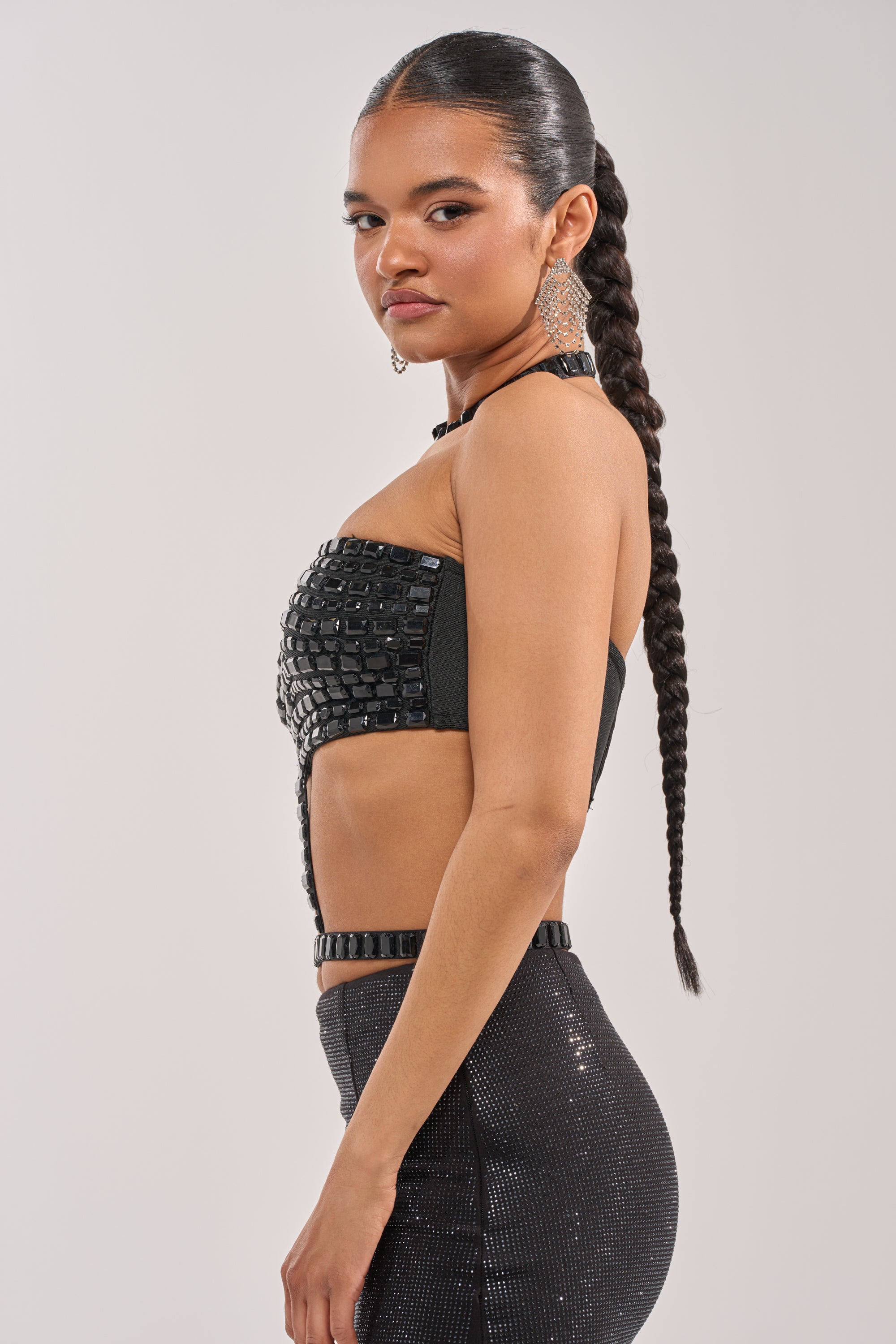 A woman with long braided hair wears the OH HOT DAMN BRALETTE and a sparkly high-waisted skirt, standing sideways and looking over her shoulder against a plain light background. 3