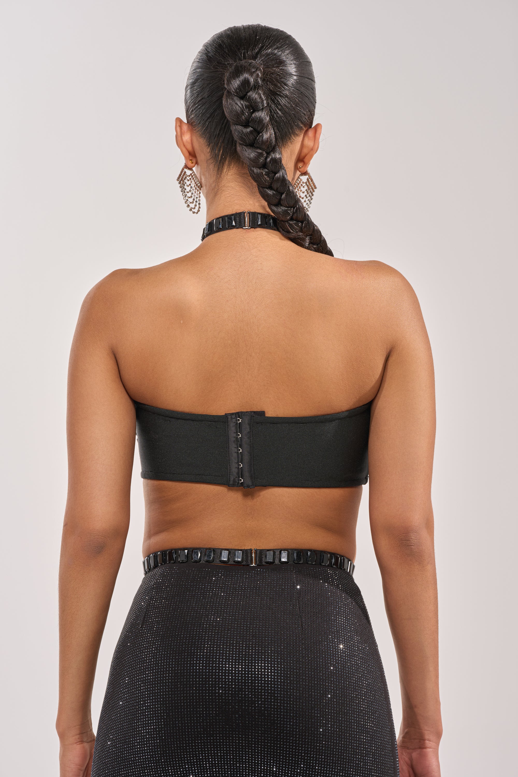 A woman with a long braid and large hoop earrings is shown from behind wearing the OH HOT DAMN BRALETTE, featuring a back clasp, paired with a high-waisted subtly sparkly black skirt, against a neutral background. 4