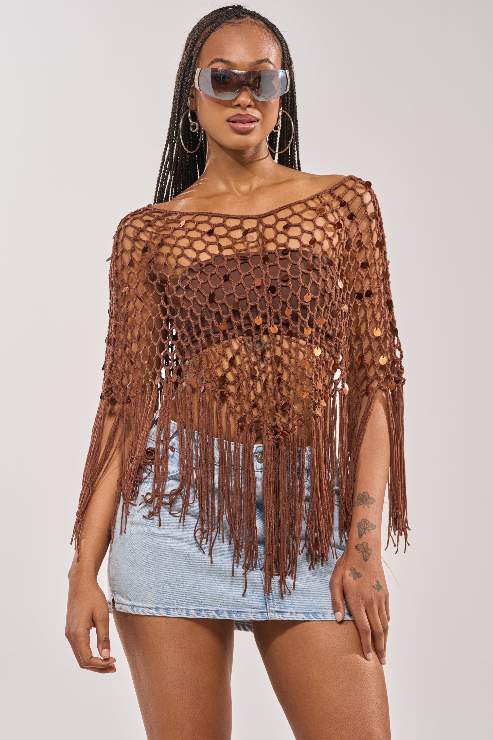 Woman in SHAKE IT QUEEN CROCHET BLOUSE over a brown bralette, large sunglasses, light blue denim mini skirt, with long braids, hoop earrings, and hand tattoos. A standout festival look against a plain background. 4