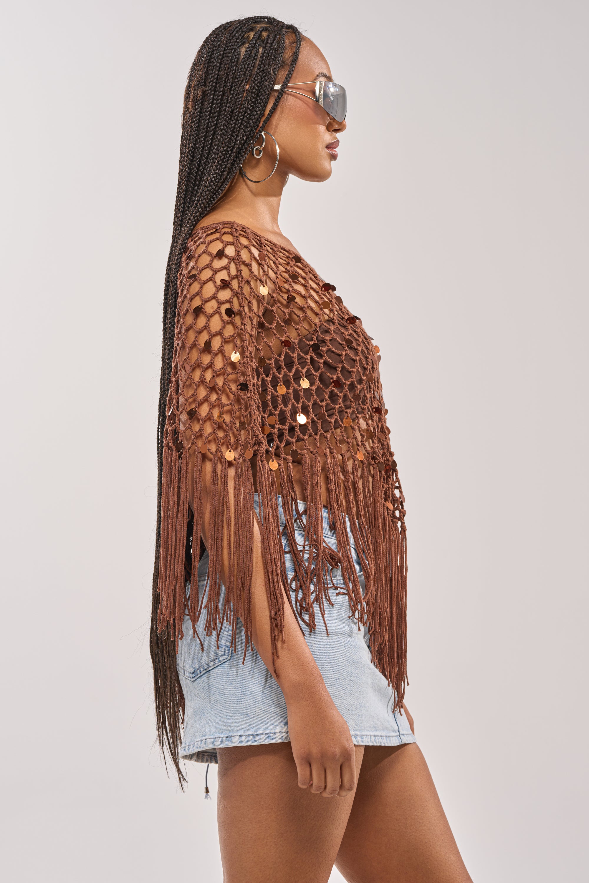 Woman in profile wearing large sunglasses, long braids, hoop earrings, and the SHAKE IT QUEEN CROCHET BLOUSE with sequins, paired with a light denim mini skirt—an ideal festival outfit against a plain background. 5