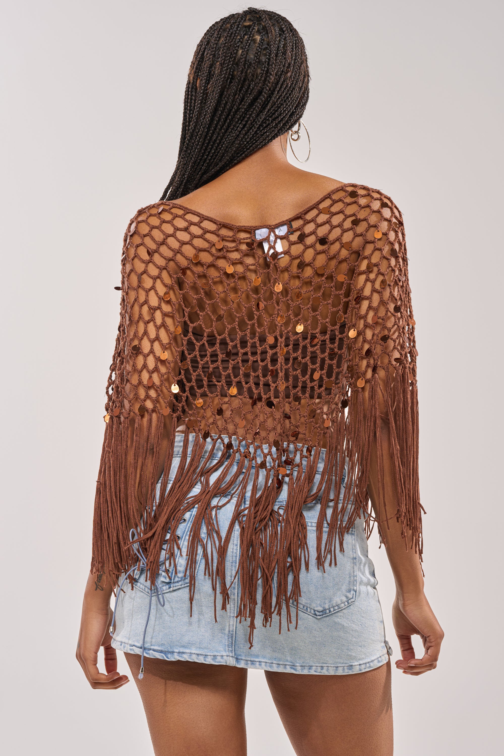 A woman with braided hair wears the SHAKE IT QUEEN CROCHET BLOUSE with fringe over a black bra and a light blue denim mini skirt, standing against a plain light background—ideal festival style. 6