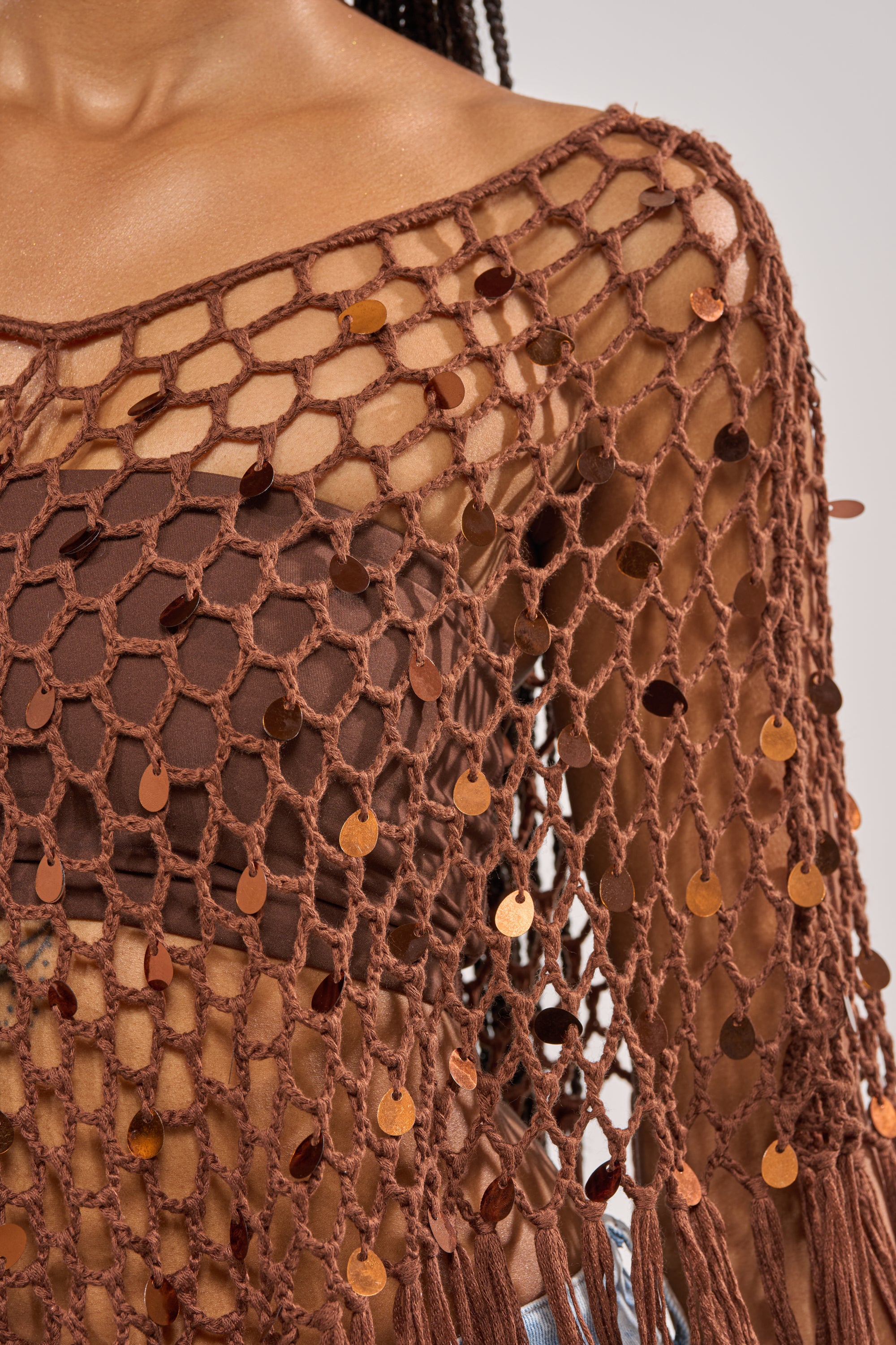 A close-up of a person in the SHAKE IT QUEEN CROCHET BLOUSE, a netted, long-sleeve cover-up with shiny sequins. This brown bandeau top combo is perfect for festivals and beautifully complements medium brown skin. 10