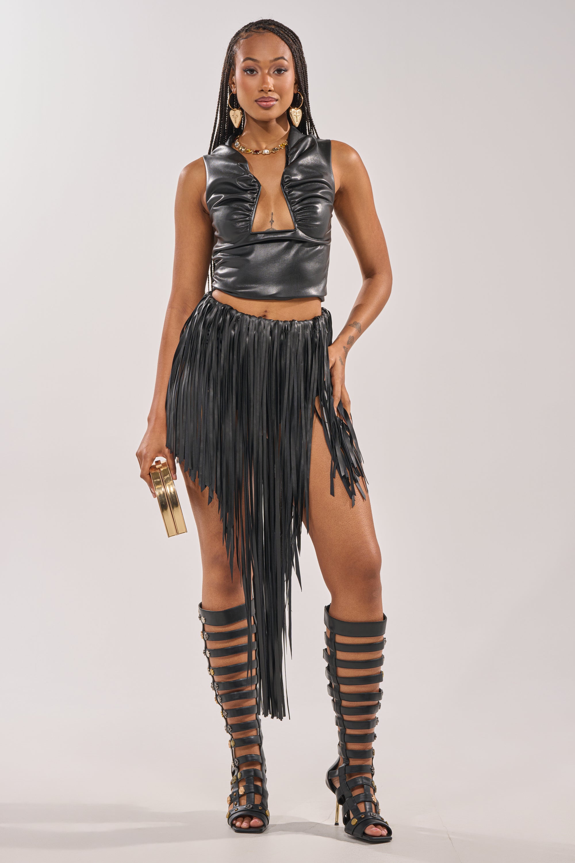 A woman stands confidently against a plain light background, wearing the COACHELLA FRINGE SKIRT, a black faux leather plunging crop top, knee-high gladiator sandals, gold earrings, and holding a gold clutch. 4