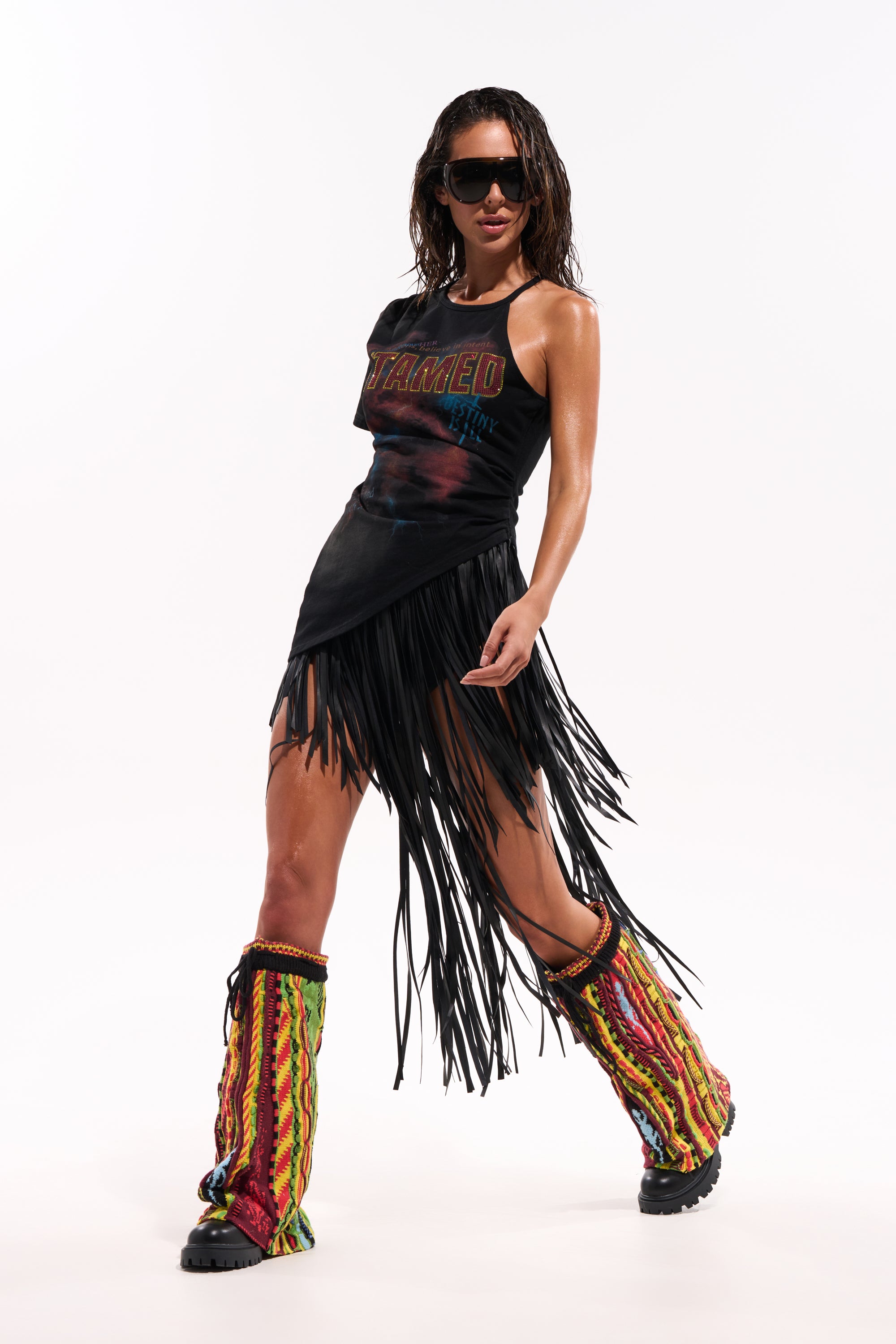 A person in sunglasses, a black sleeveless "TAMED" top, the COACHELLA FRINGE SKIRT, and colorful crochet leg warmers over black boots poses confidently against a plain white background. 1