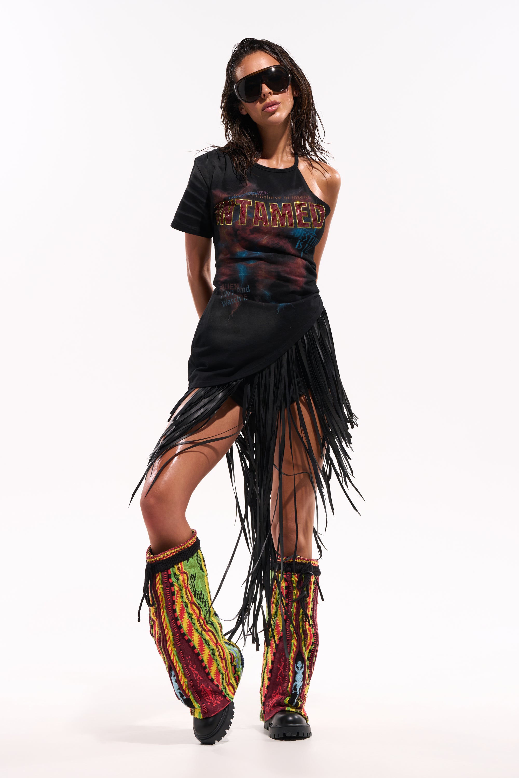 Wearing dark sunglasses, a black "UNTAMED" tee, the COACHELLA FRINGE SKIRT, and colorful knee-high boots, a person stands confidently against a plain white background. 2