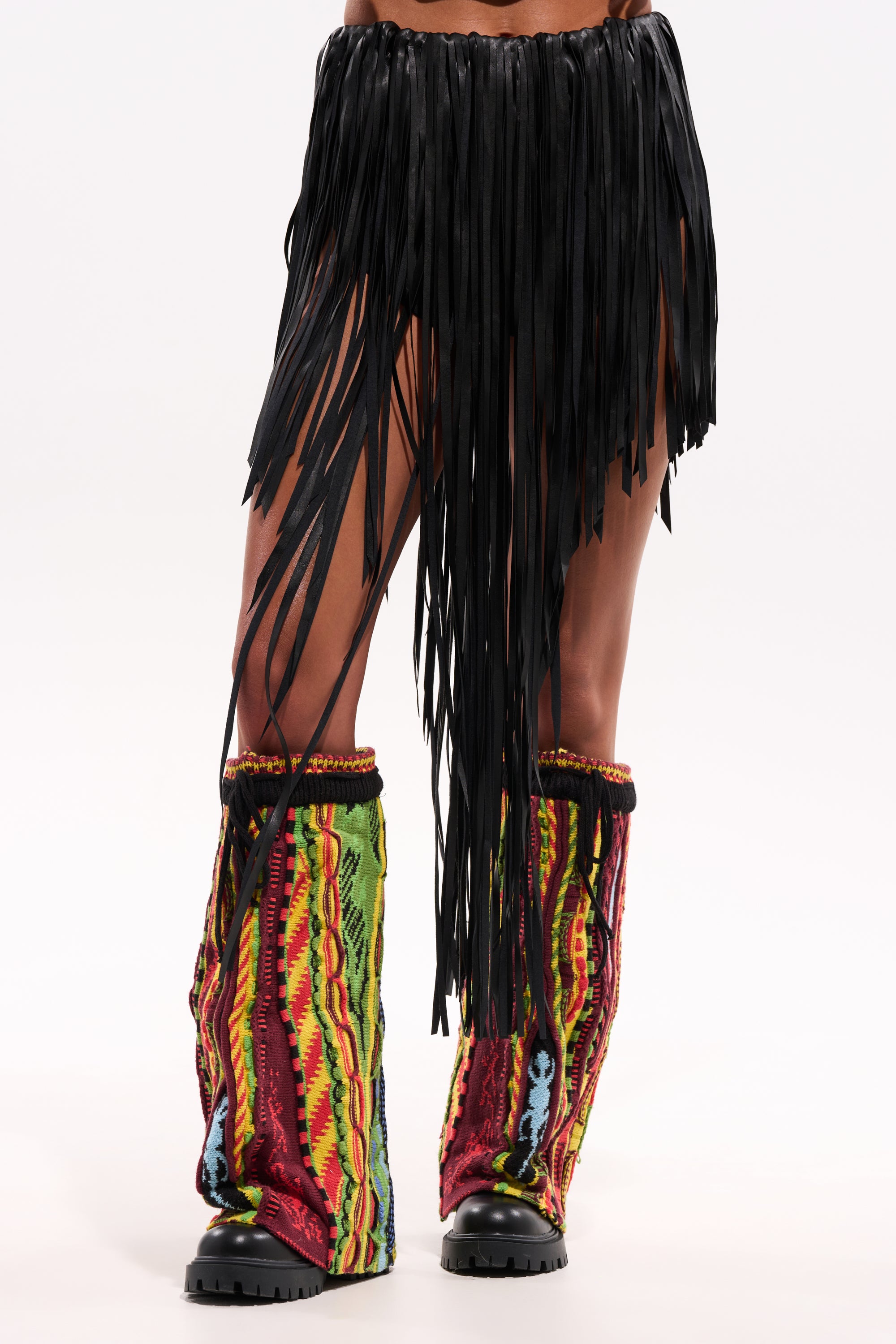 Person wearing the COACHELLA FRINGE SKIRT with colorful patterned leg warmers over black boots, standing against a plain white background. 3