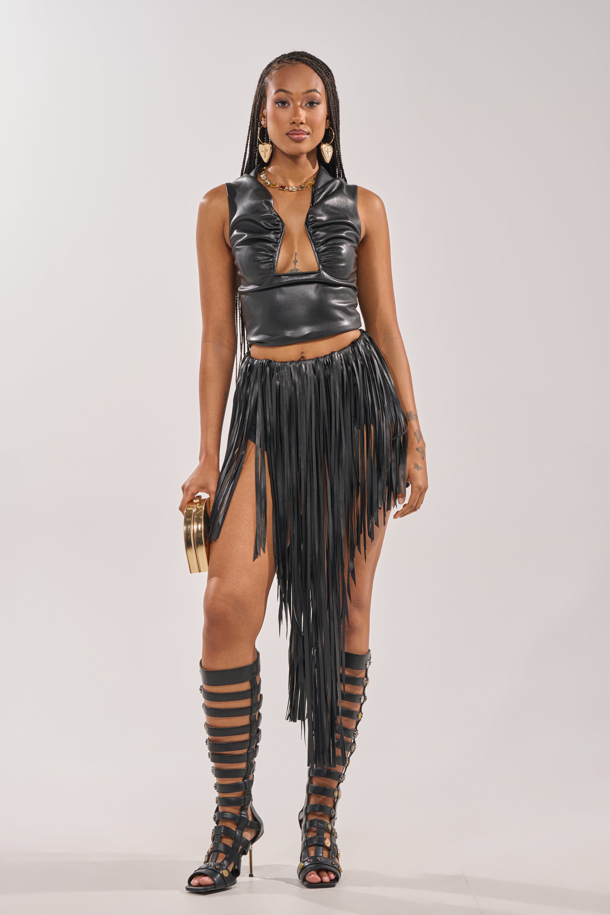 A woman stands confidently in a black leather sleeveless crop top with a front cutout, the COACHELLA FRINGE SKIRT, gladiator-style knee-high sandals, gold earrings, and holding a small round clutch. 8