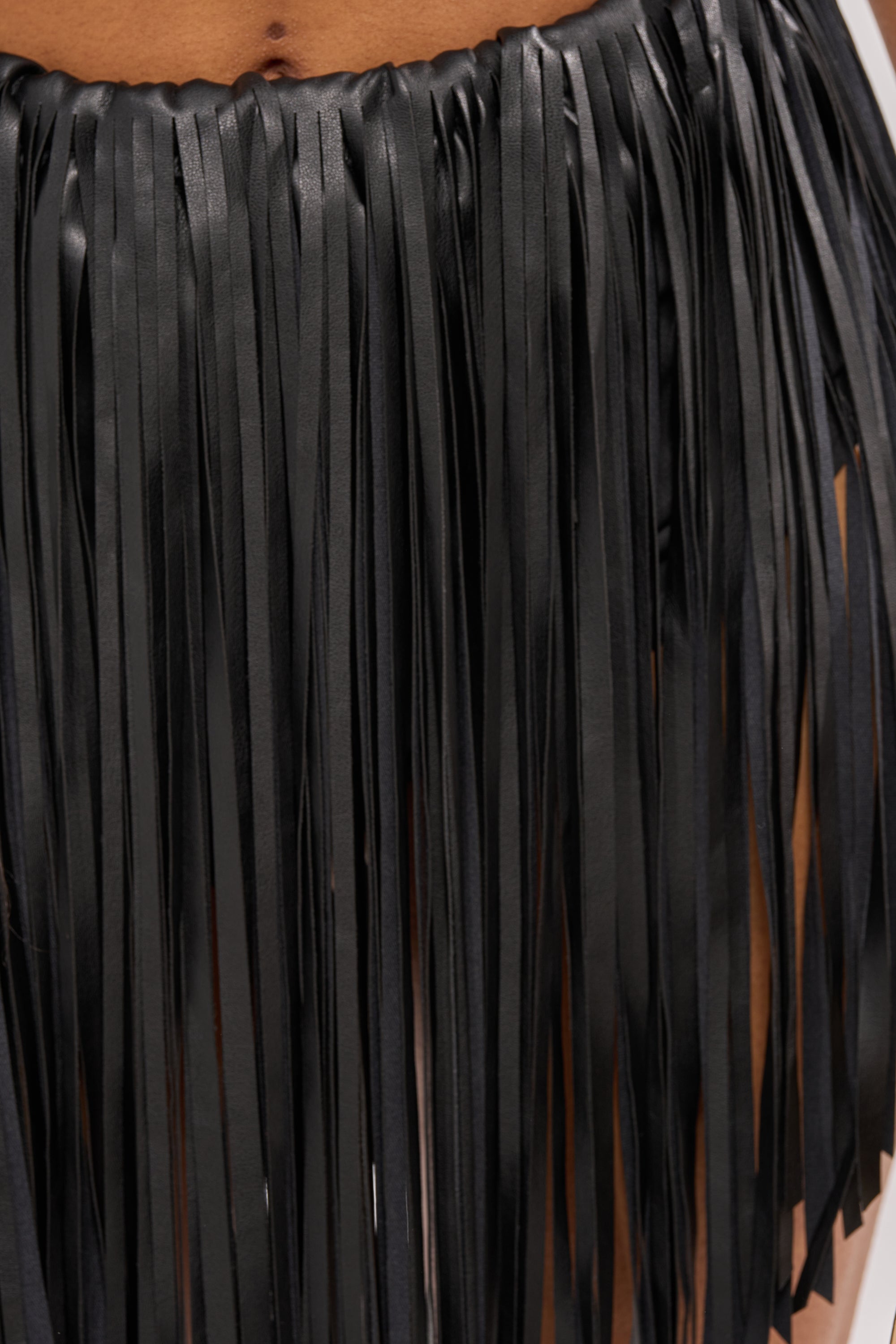 Close-up of a person wearing the COACHELLA FRINGE SKIRT—black faux leather with long, thin fringe strips that reveal glimpses of skin underneath. 10