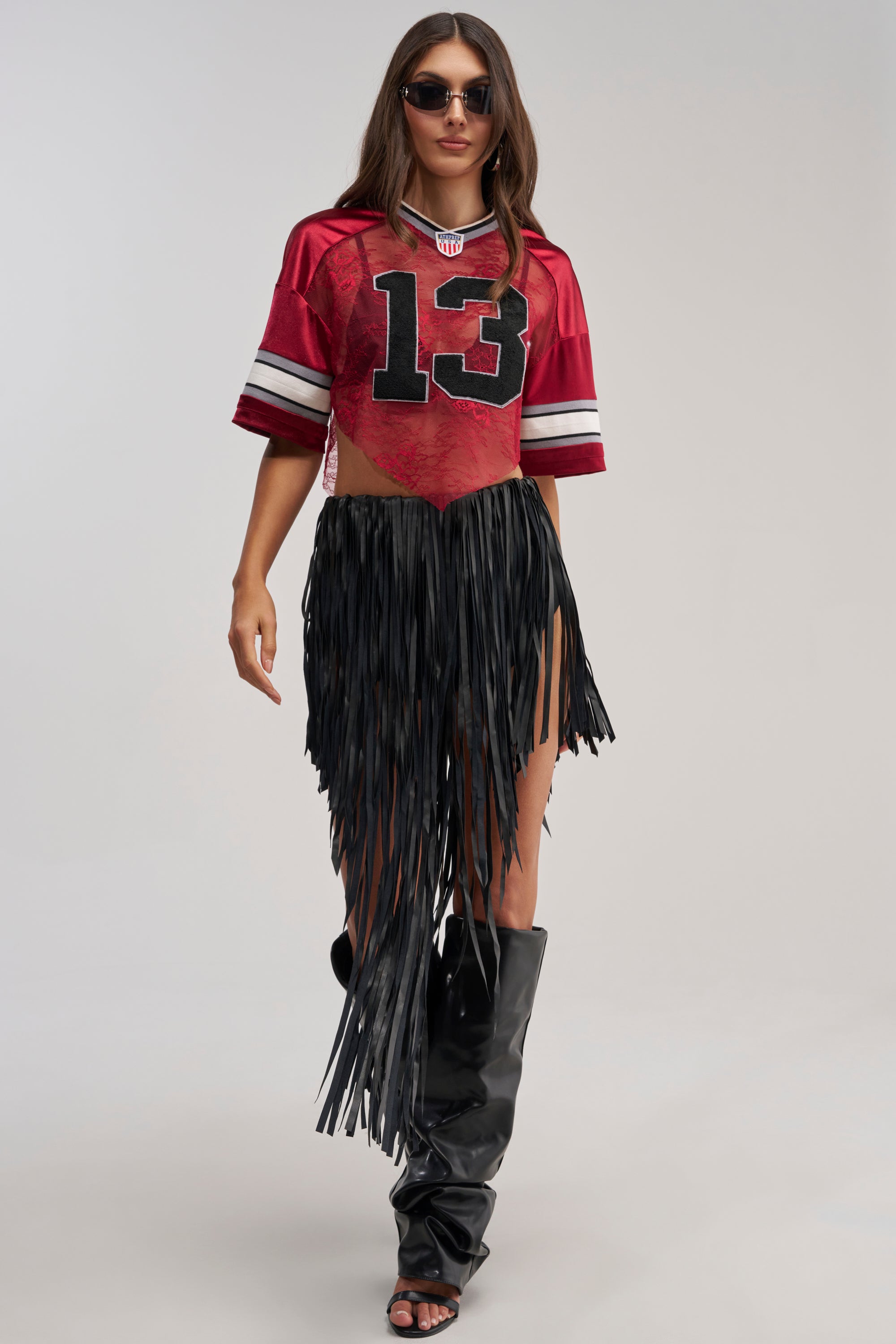 A woman confidently poses against a plain light background wearing sunglasses, a red football jersey with the number 13, tall black boots, and the COACHELLA FRINGE SKIRT.