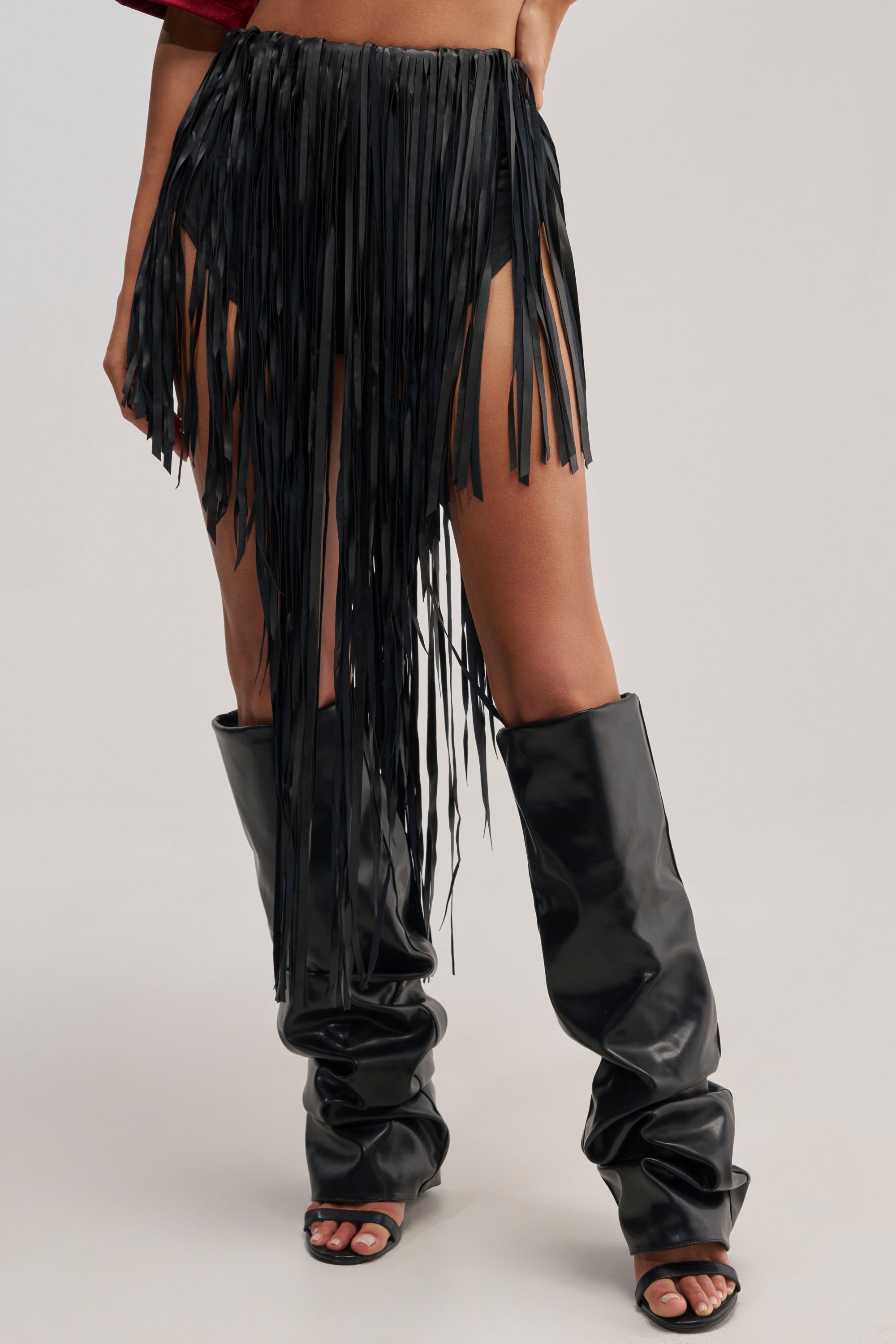 Wearing the COACHELLA FRINGE SKIRT, styled with black slouchy knee-high boots and high-heeled sandals, the look is bold and festival-ready against a plain, light-colored backdrop.