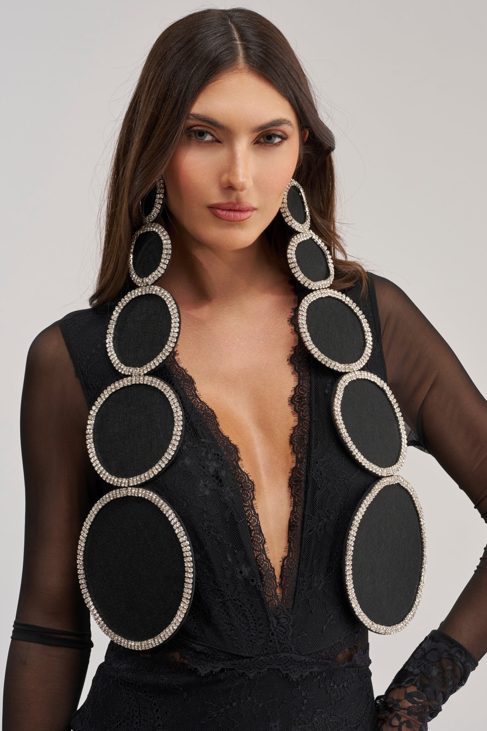 A woman with long brown hair wears a sheer black lace top and TO THE DISCO EARRING—sparkling rhinestone statement earrings with black centers—gazing confidently at the camera against a neutral background.