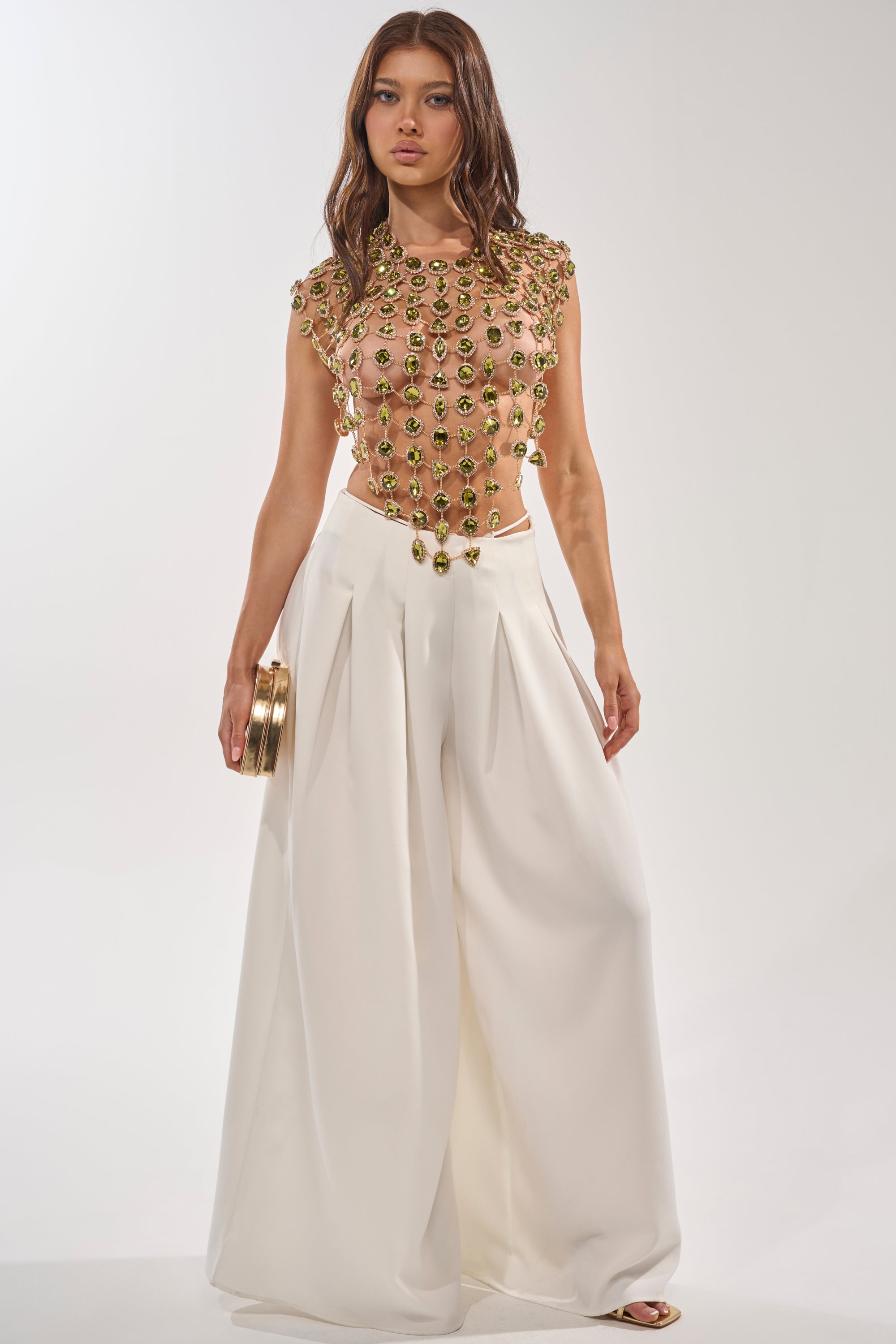 Woman stands against a white background wearing the ALL I NEED IS MONEY RHINESTONE CHAIN TOP—a glamorous, sheer, sleeveless top with emerald rhinestone details—paired with high-waisted wide-leg white trousers. She holds a gold clutch and has loose, wavy hair. 5