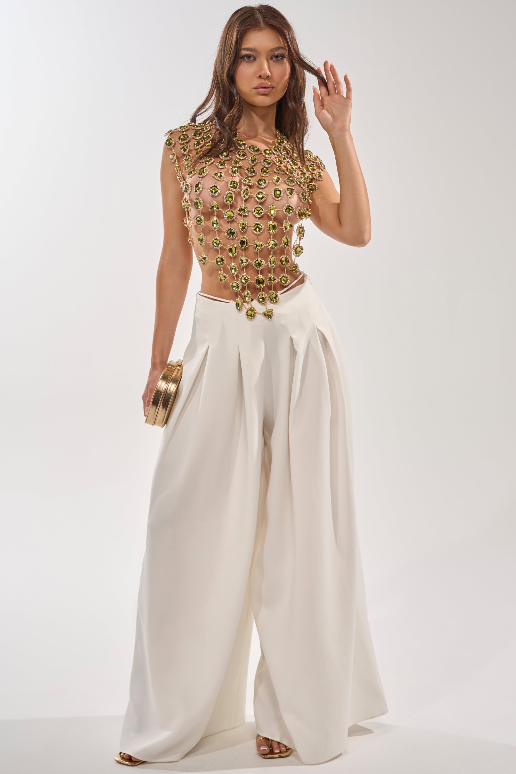 A woman models the ALL I NEED IS MONEY RHINESTONE CHAIN TOP, decorated with gold and green accents, paired with wide-leg white pants, a small gold clutch, and gold heels against a plain white background. 6