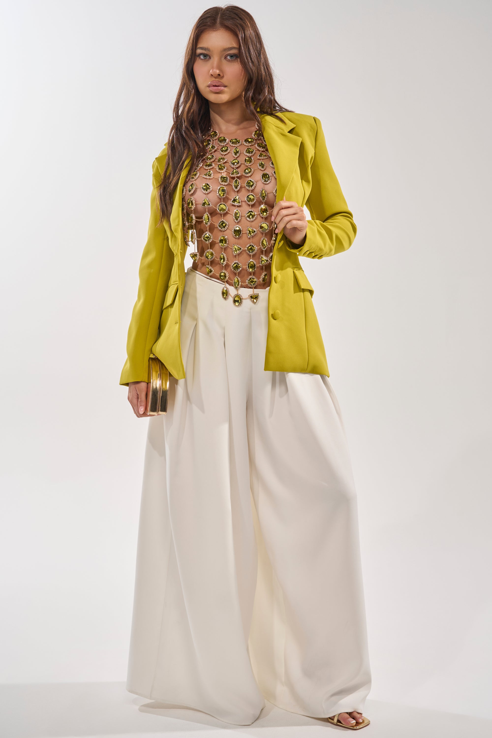 A woman stands against a plain background wearing the ALL I NEED IS MONEY RHINESTONE CHAIN TOP with a yellow blazer and wide-leg white pants. She holds a clutch, her long hair loose, exuding confidence and modern style. 7